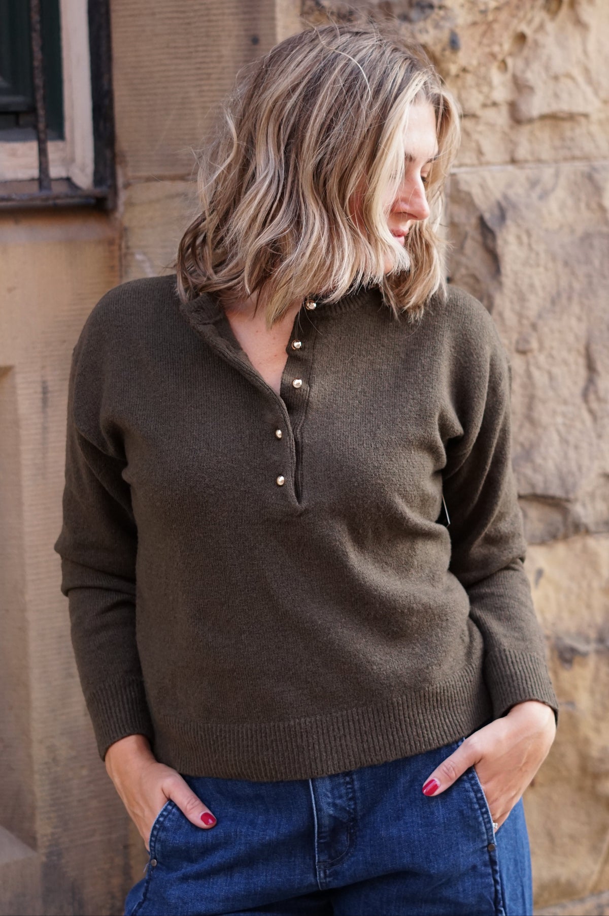 A woman wearing a dark brown ribbed knit henley sweater with a buttoned placket, paired with blue jeans, standing against a rustic stone wall.