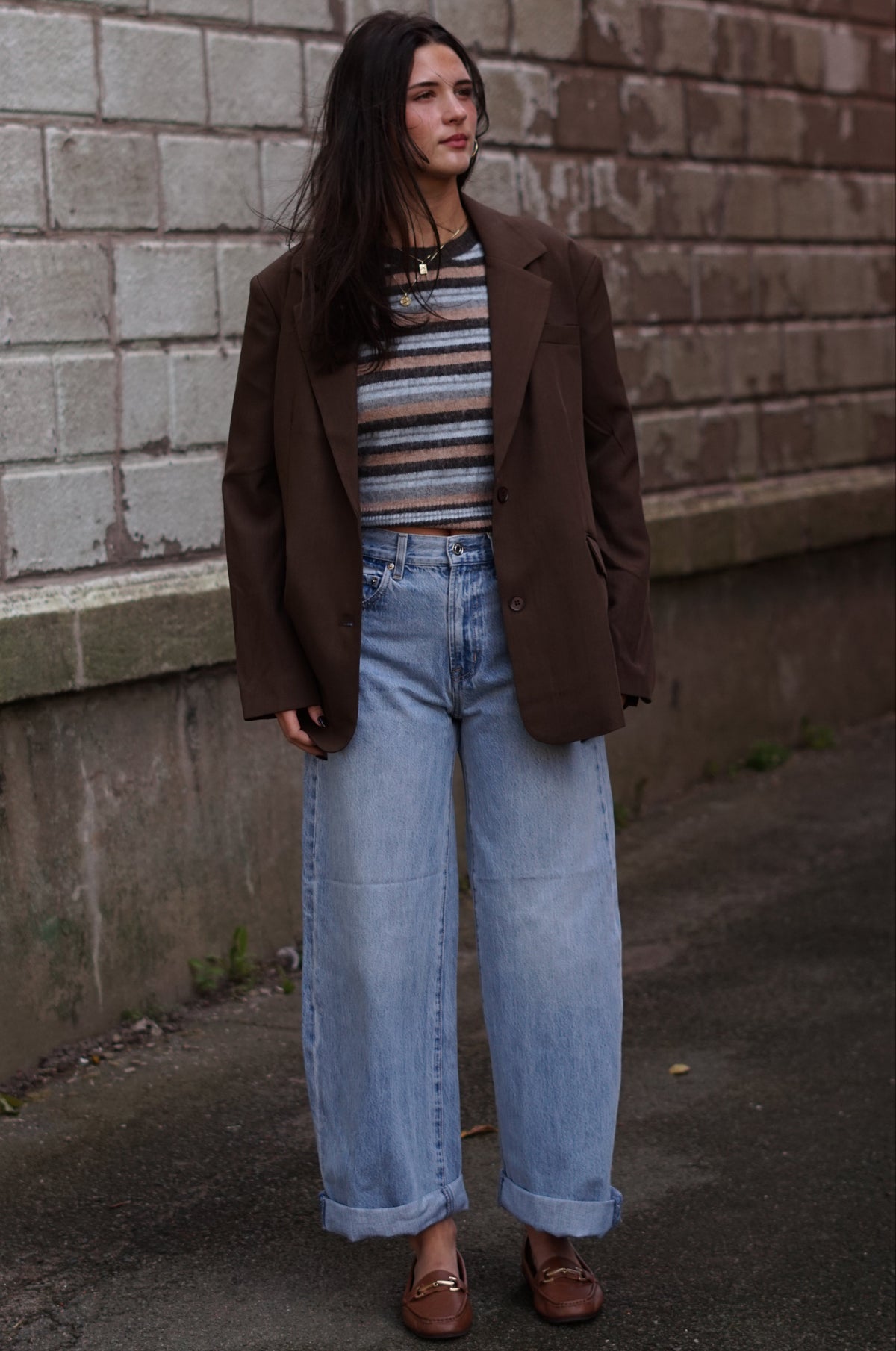 A woman stands outdoors against a weathered brick wall wearing a brown blazer, a striped knit top, light-wash wide-leg jeans, and brown leather loafers.