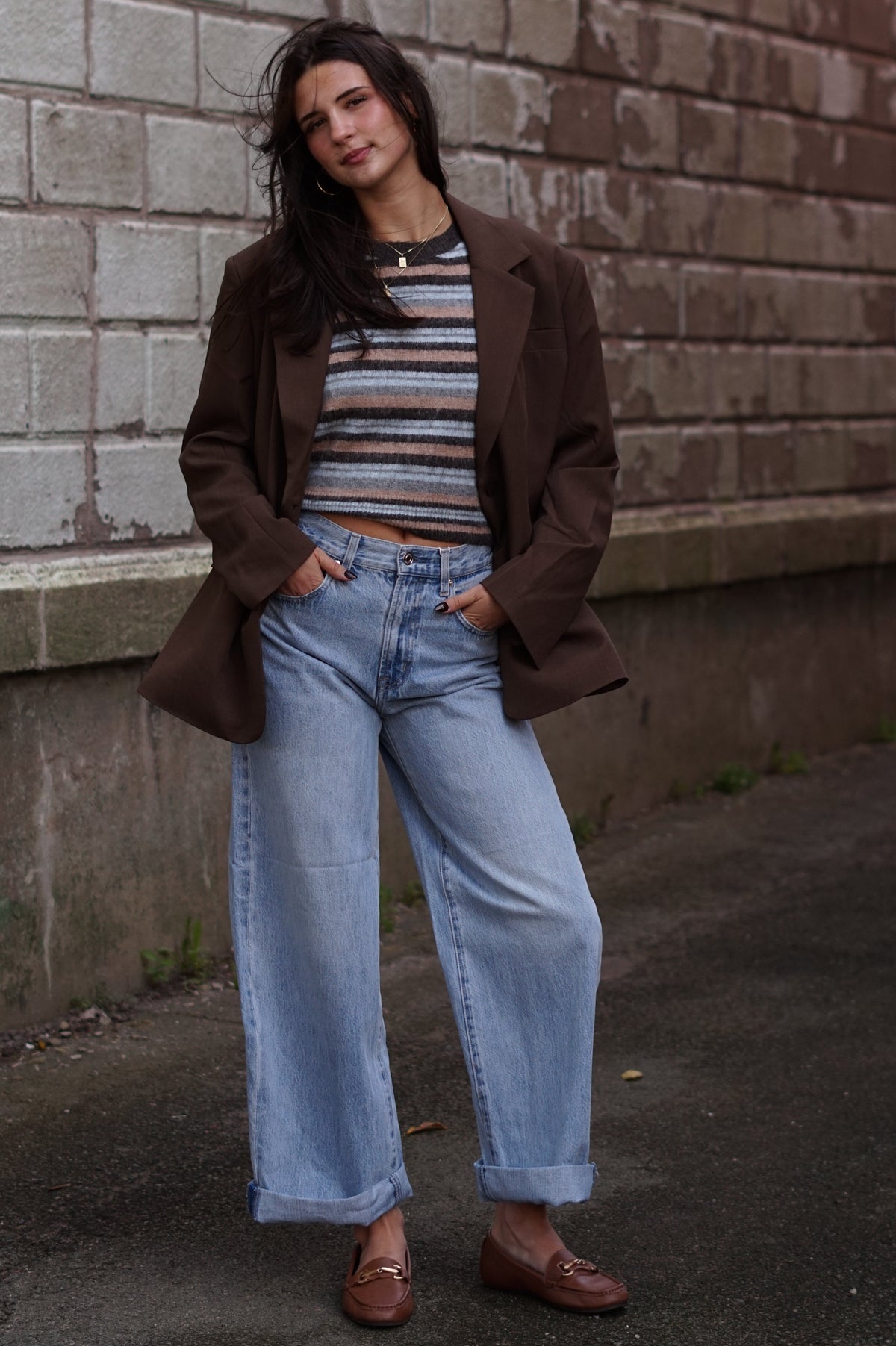 A woman stands outdoors against a brick wall wearing an oversized brown blazer over a striped crop top, light blue wide-leg jeans, and brown loafers.