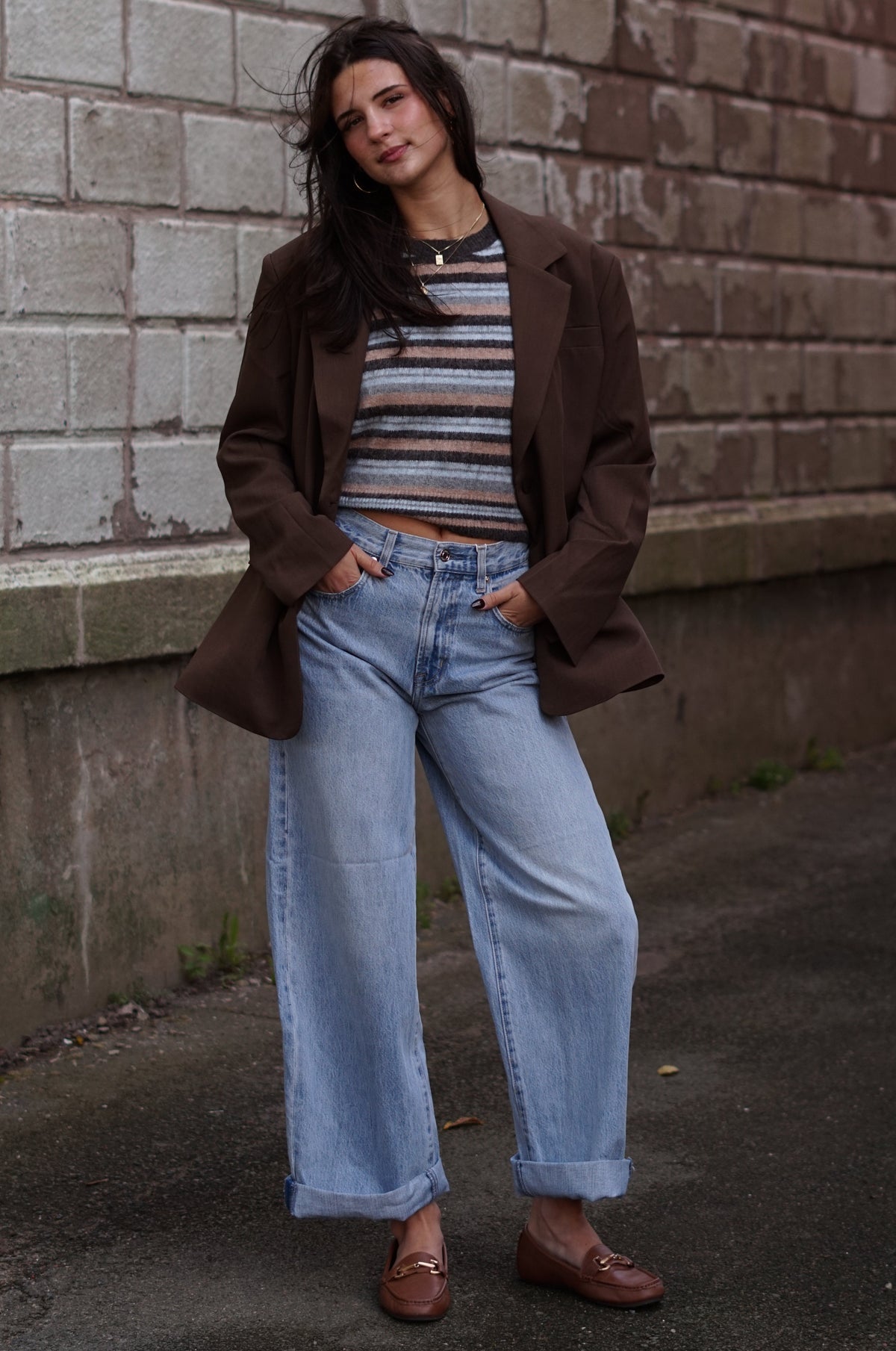 A woman stands outdoors against a brick wall wearing a brown blazer, a striped knit top, light blue wide-leg jeans, and brown loafers.