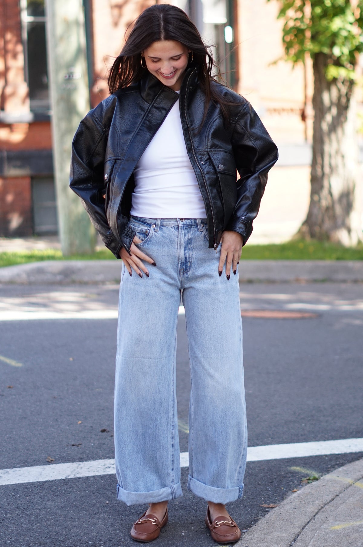 A woman stands on a city street wearing a black leather bomber jacket, a white ribbed tank top, wide-leg light blue jeans, and brown leather loafers.