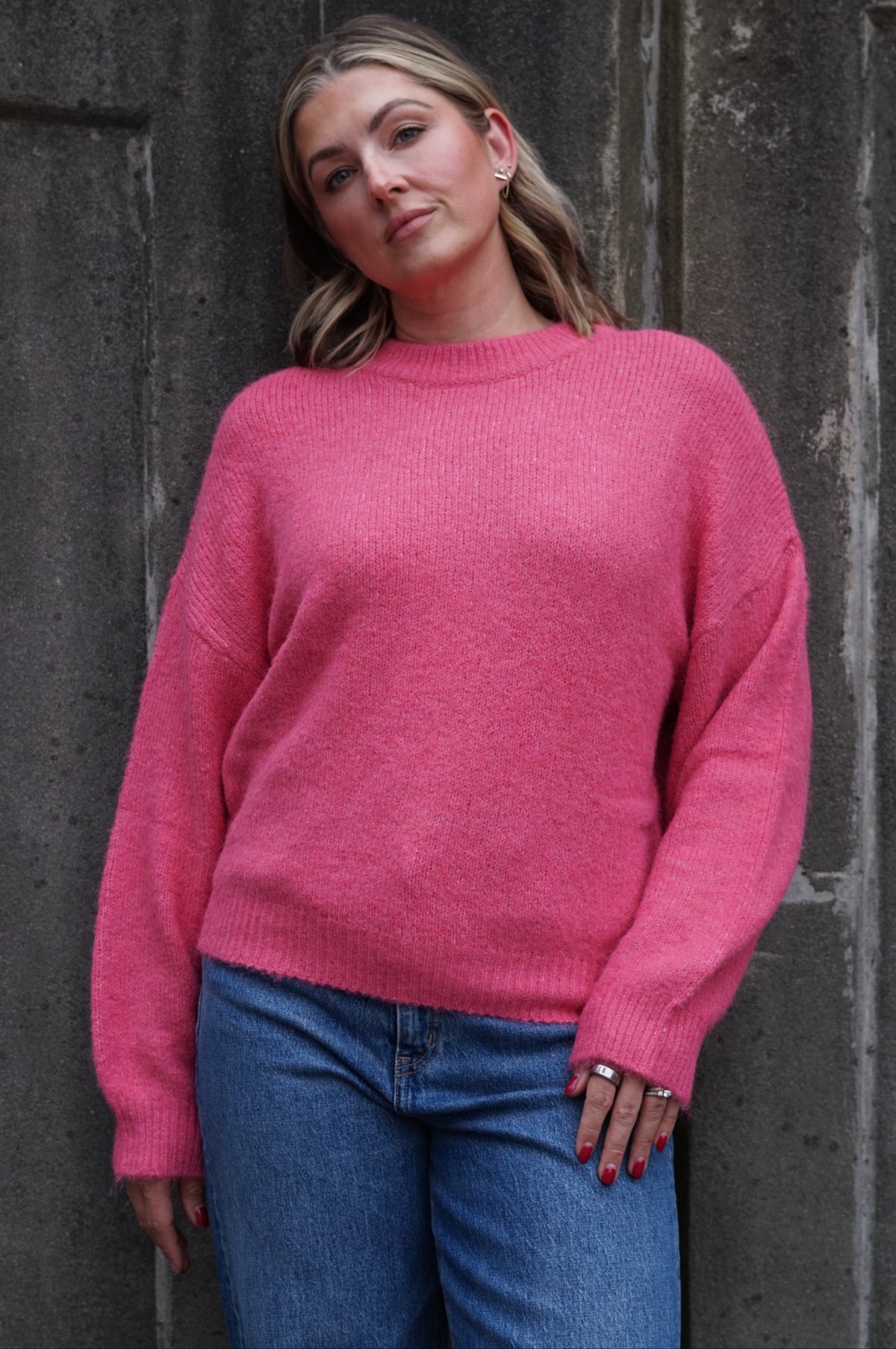 A woman wearing a bright pink knit crewneck sweater and blue jeans, standing against a dark textured stone wall.
