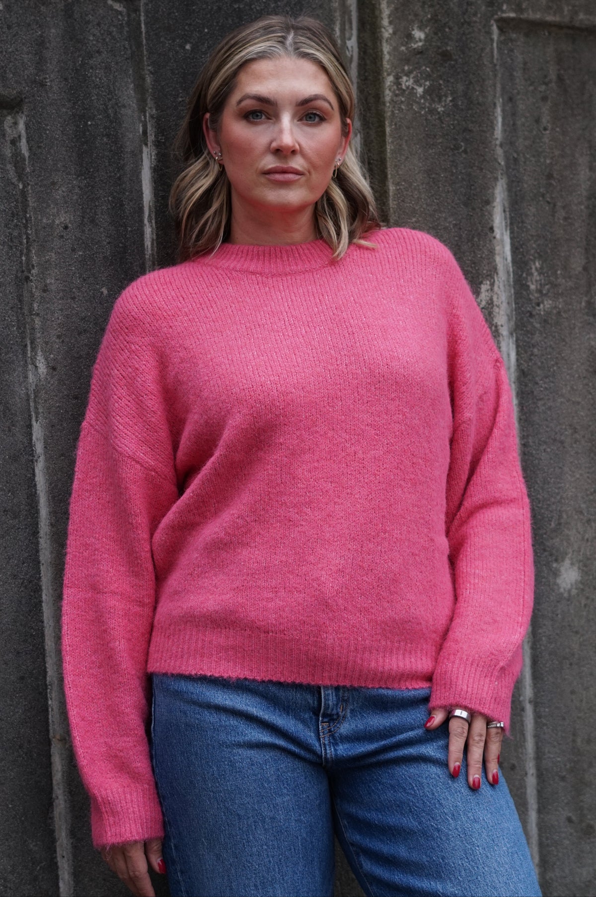 A woman wearing a pink knit crewneck sweater and blue jeans, standing against a dark textured stone wall.