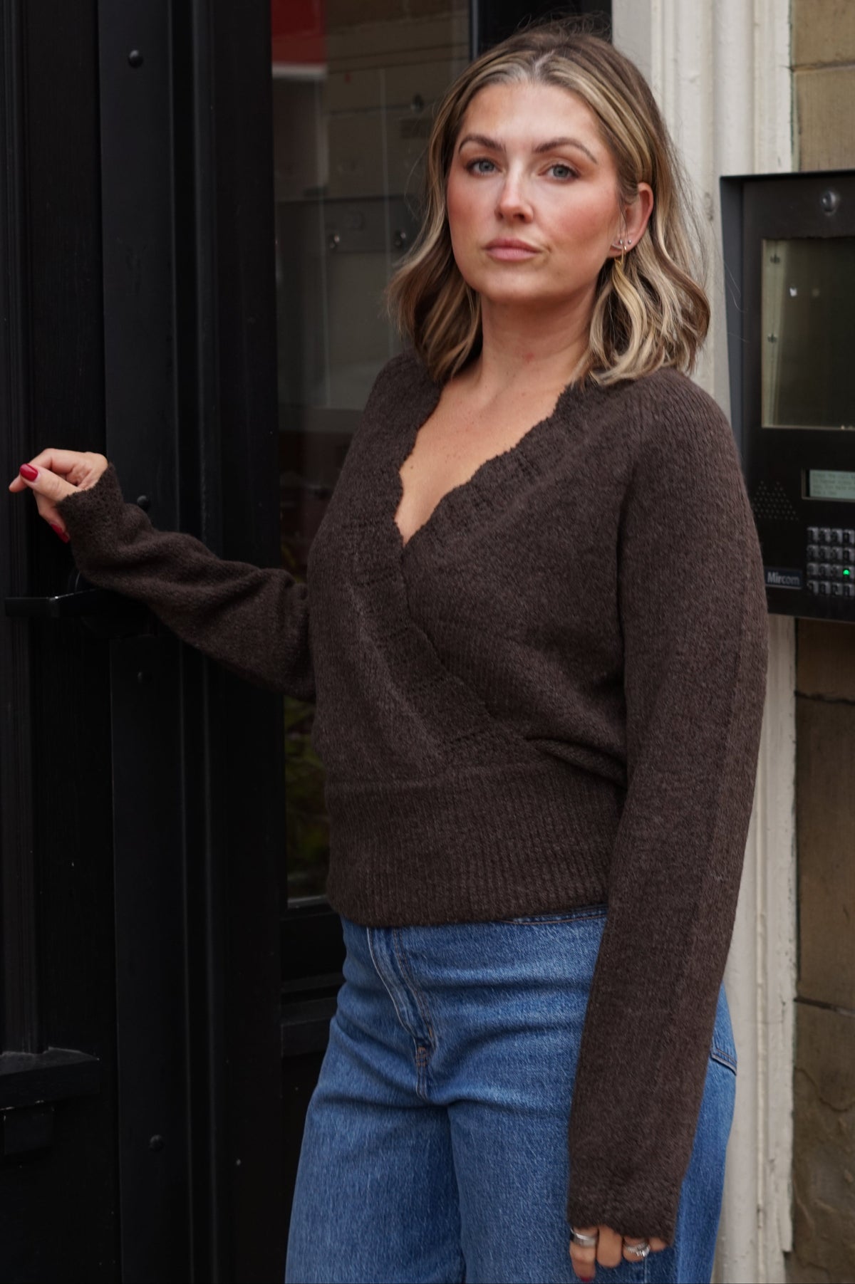 A woman wearing a dark brown knit V-neck sweater and blue jeans, standing next to a black door.