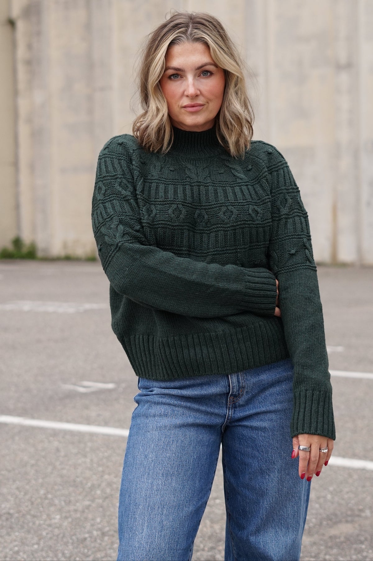 A woman wearing a dark forest green cable-knit mock neck sweater and blue jeans, standing outdoors in front of a concrete wall.