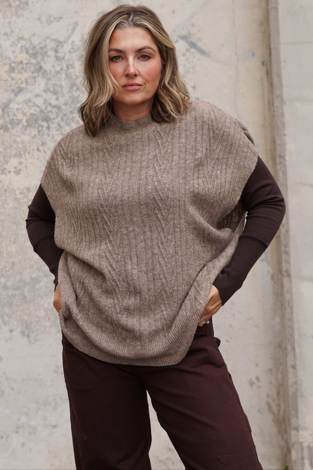 A woman wearing a chunky cable-knit poncho sweater in taupe over a dark brown long-sleeve shirt and dark brown pants, standing against a textured concrete wall.