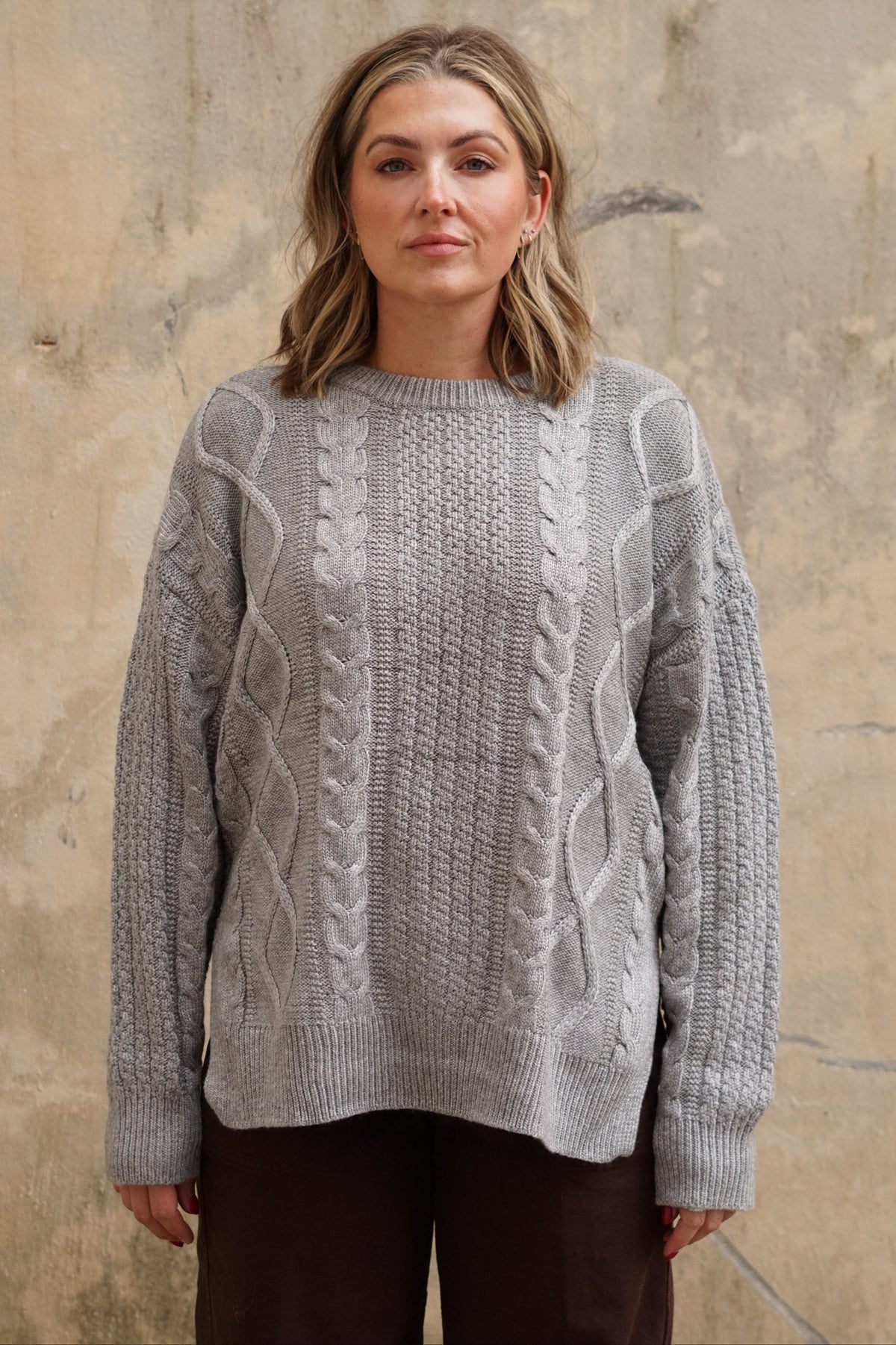 A woman wearing a light gray cable-knit sweater and dark brown pants, standing against a textured beige wall.