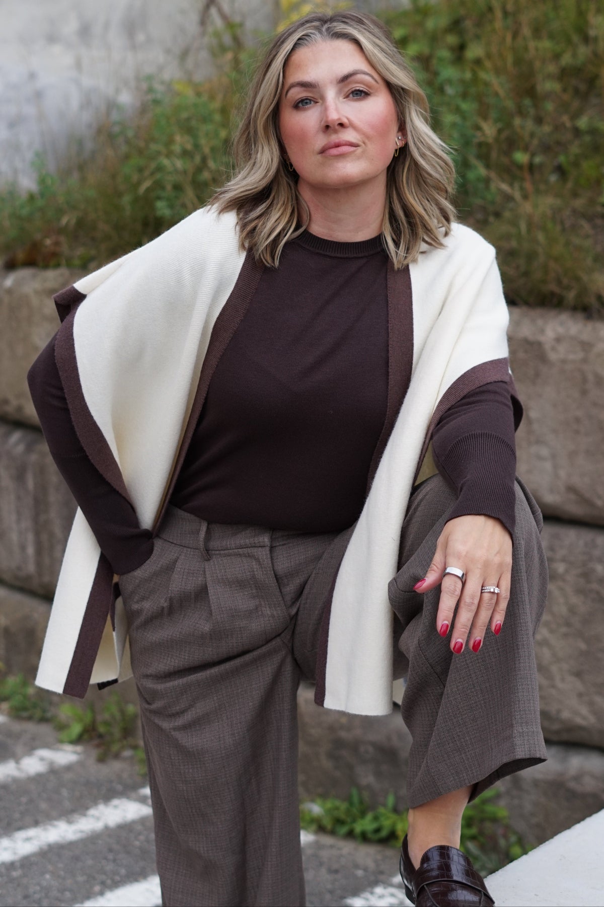 A woman posing outdoors in a stylish outfit featuring a dark brown long-sleeve top, taupe wide-leg trousers, and a cream wrap jacket with dark brown trim.