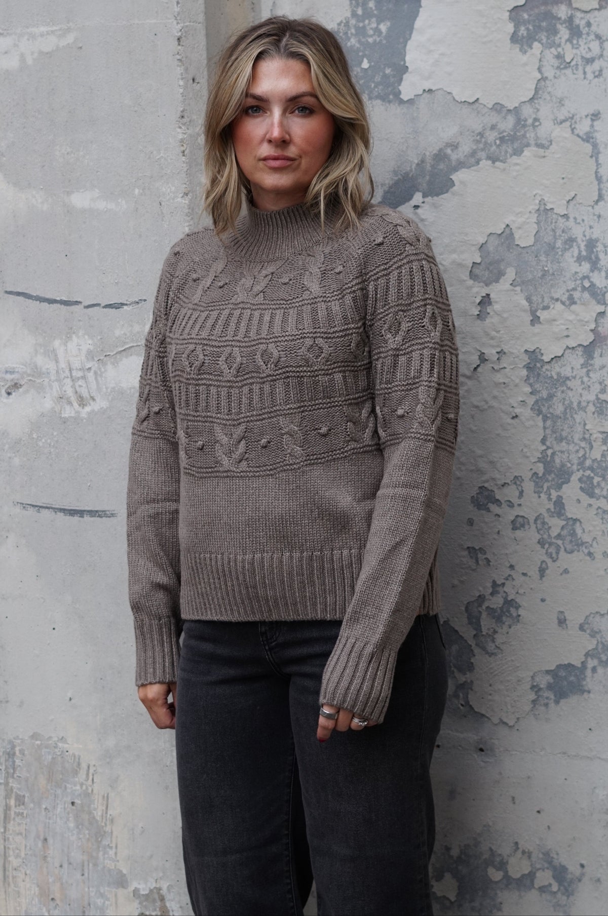 A woman wearing a taupe cable-knit mock-neck sweater and dark jeans, standing against a textured, weathered gray concrete wall.