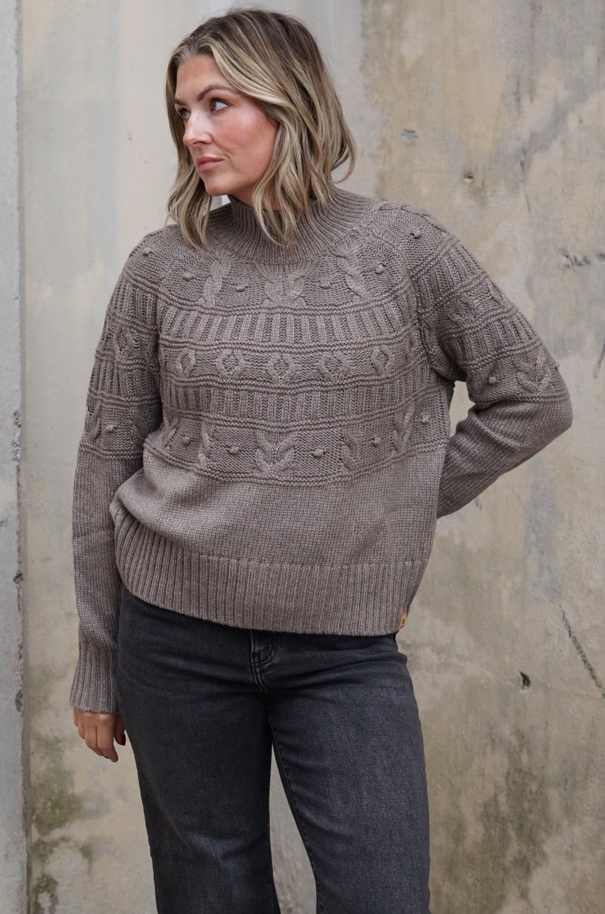 A woman wearing a taupe-brown cable-knit sweater with geometric patterns on the chest, paired with dark gray jeans, standing against a textured concrete wall.