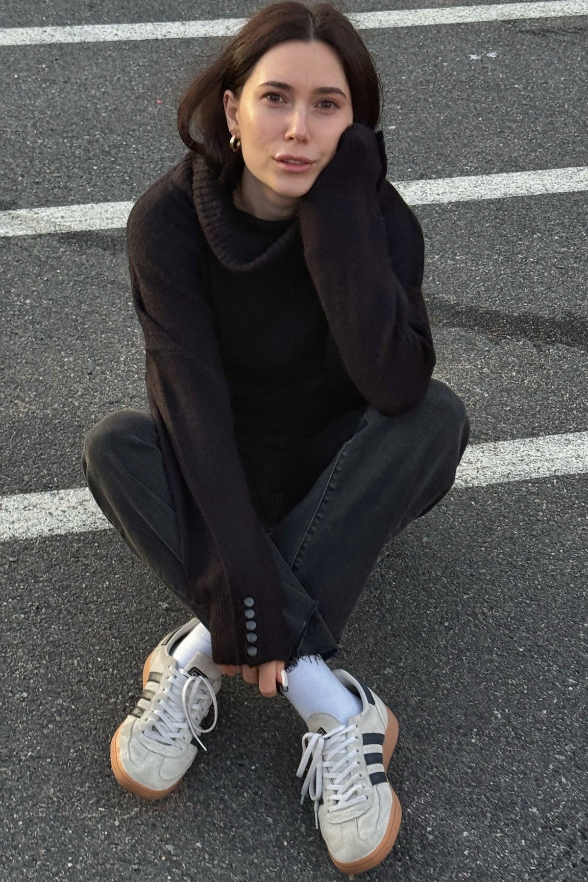A woman crouching in a parking lot wearing a black turtleneck sweater, black wide-leg jeans, white socks, and white sneakers with black stripes.