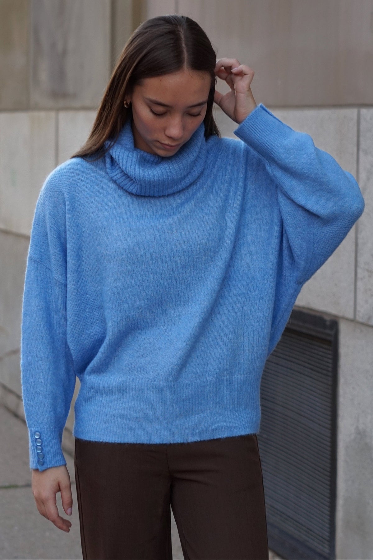 A woman wearing a light blue knit turtleneck sweater with wide batwing sleeves and dark brown pants, standing outdoors against a stone wall.