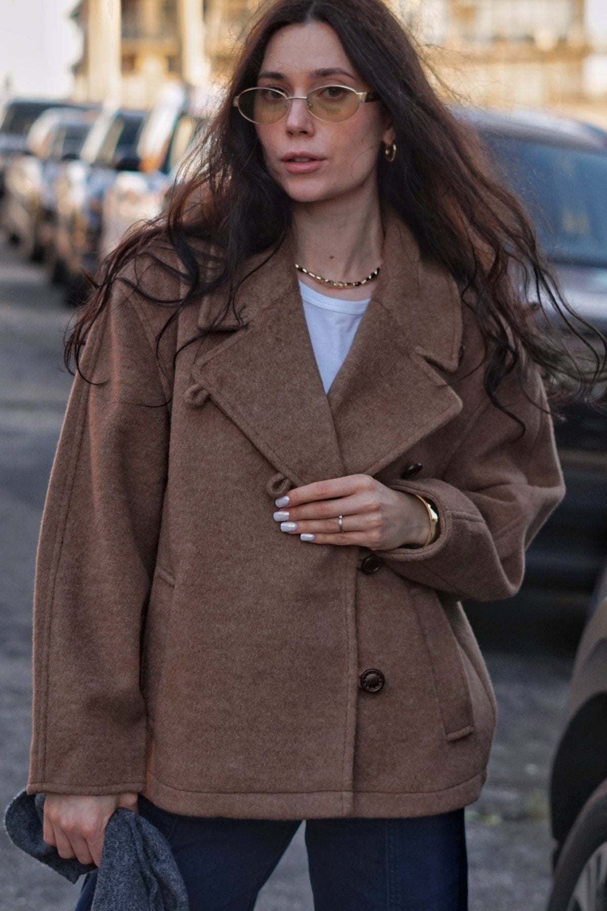 A woman wearing a brown double-breasted coat over a white top and dark blue jeans, standing on a city street with parked cars in the background.