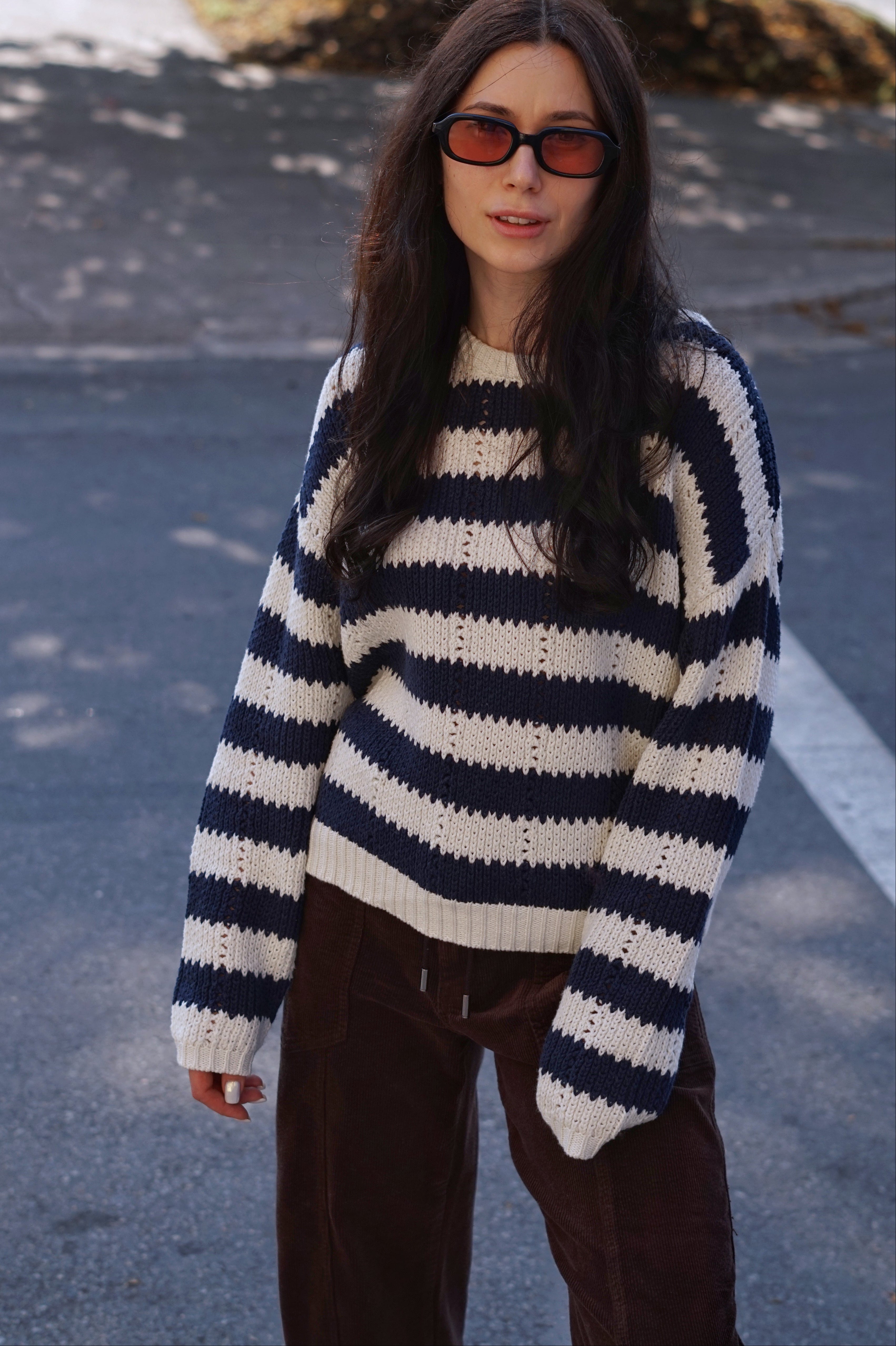 A woman wearing a navy and cream striped knit sweater, dark brown corduroy pants, and black sunglasses with orange-tinted lenses, standing outdoors on a paved street.