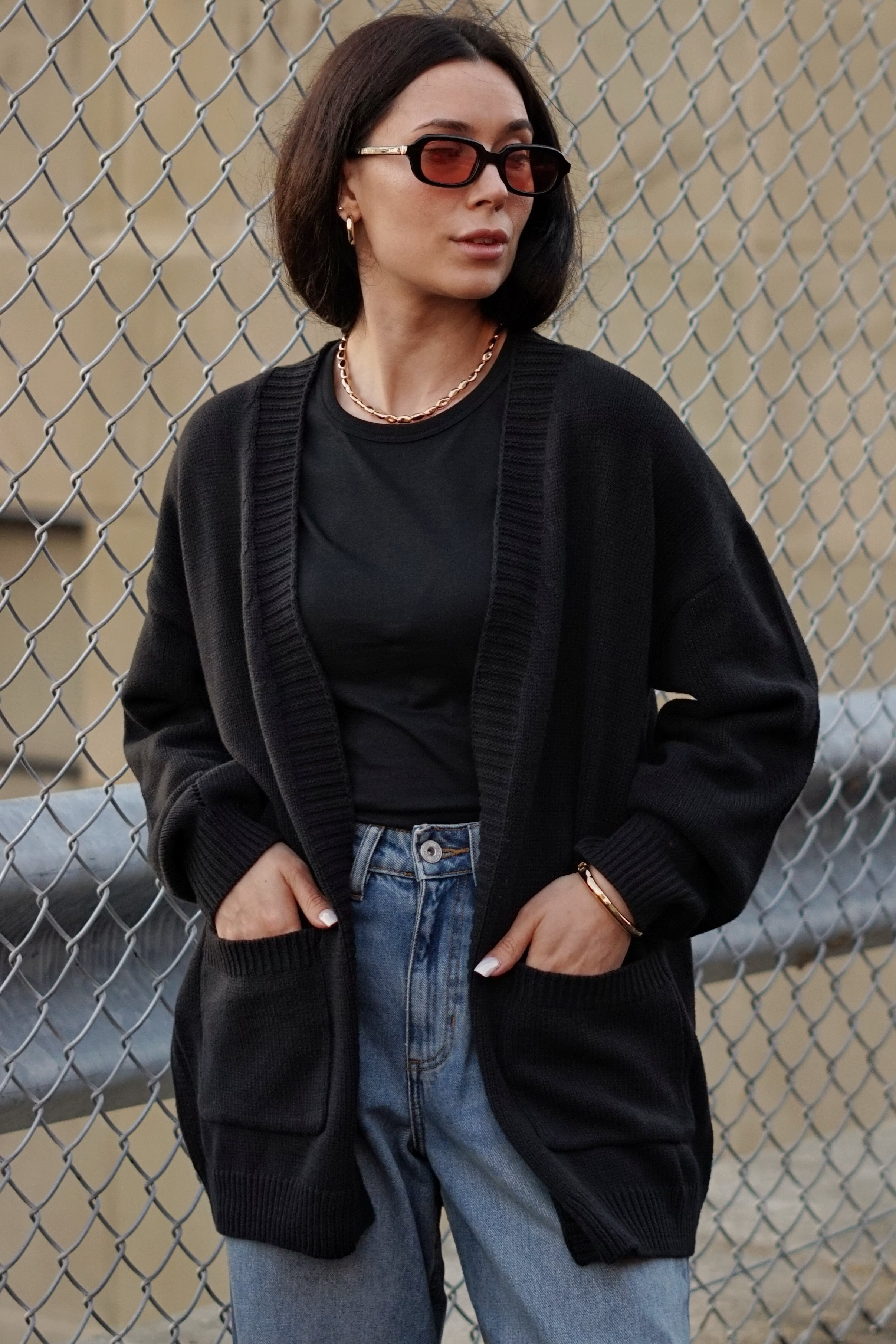A woman with short dark hair wearing a black knit cardigan over a black top, blue jeans, and red-framed sunglasses, standing in front of a chain-link fence.