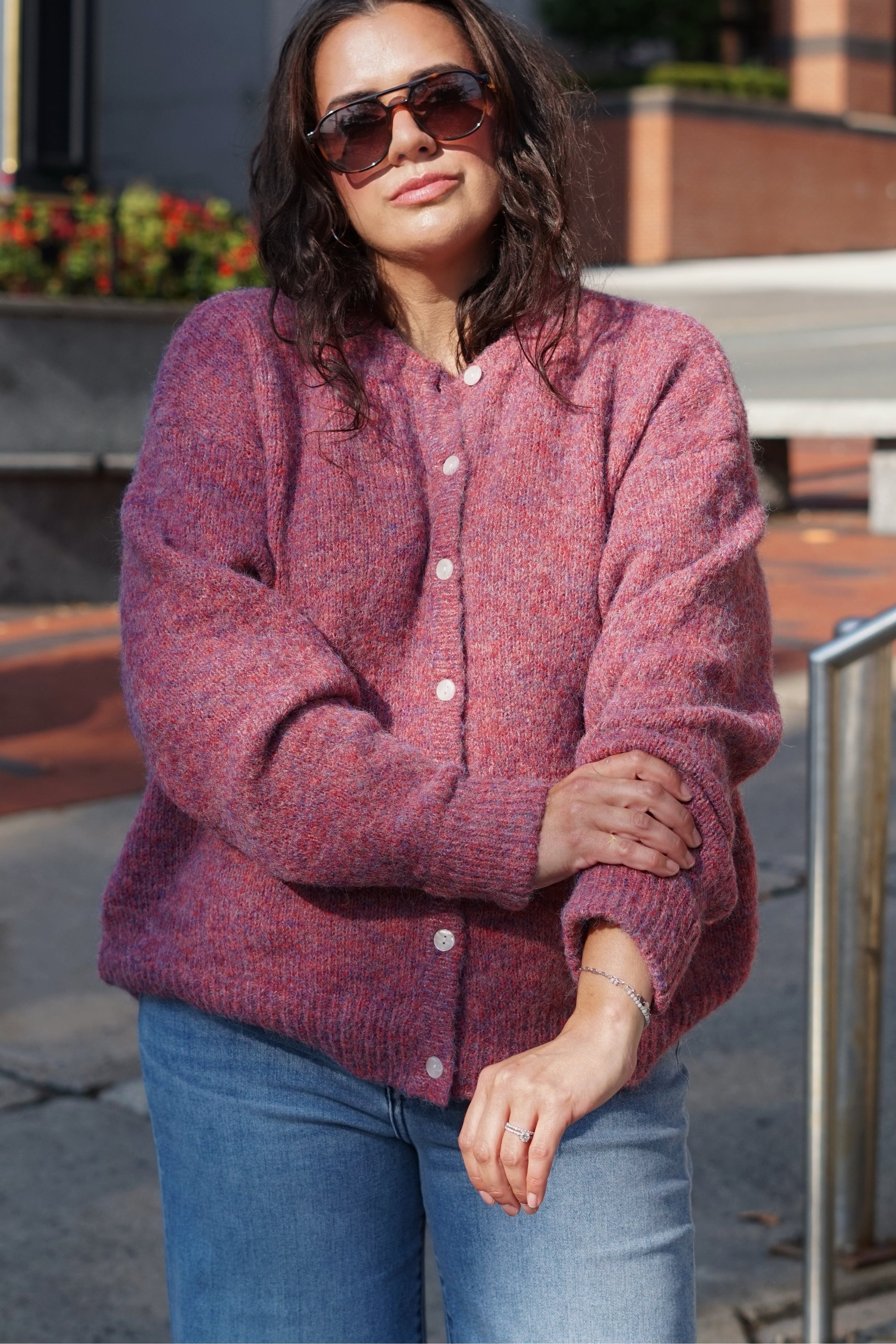 A woman wearing a marled dusty rose knit cardigan and blue jeans, standing outdoors on a city sidewalk in front of a building.