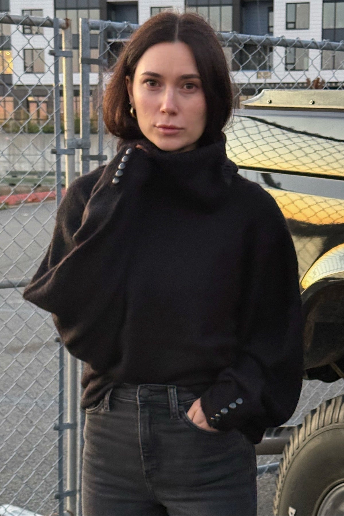 A woman wearing a black turtleneck sweater and dark gray jeans stands outdoors next to a black and gold lifted truck, with a chain-link fence and modern apartment buildings in the background.