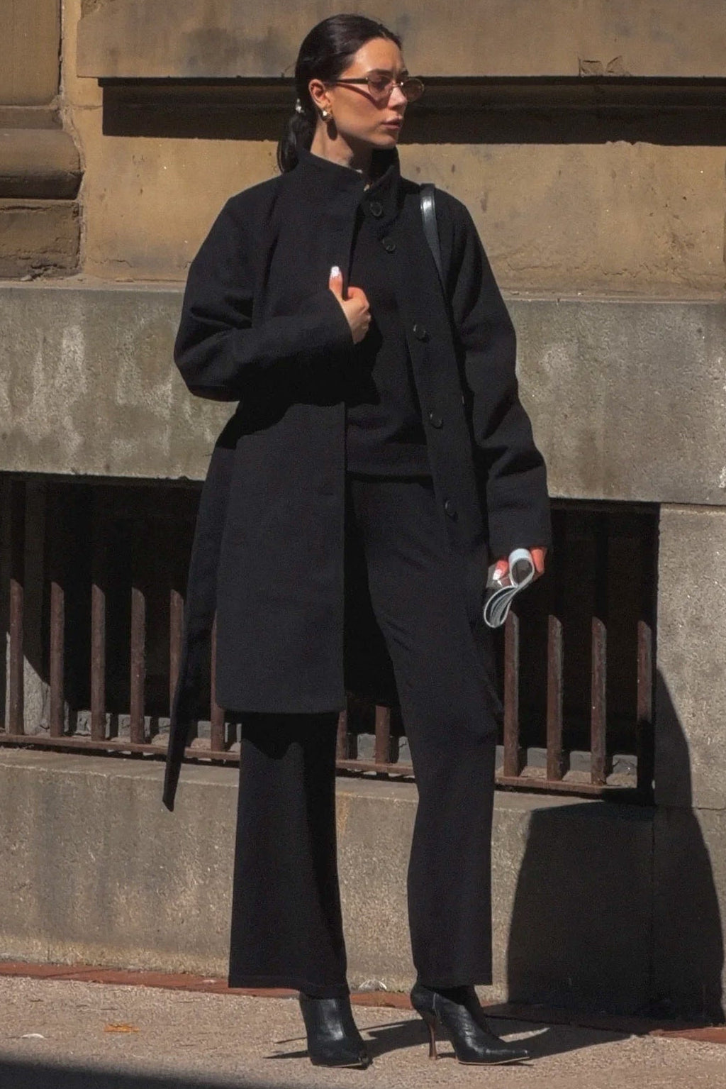 A woman wearing a black long coat, black pants, and black boots stands on a city sidewalk in front of a stone building.