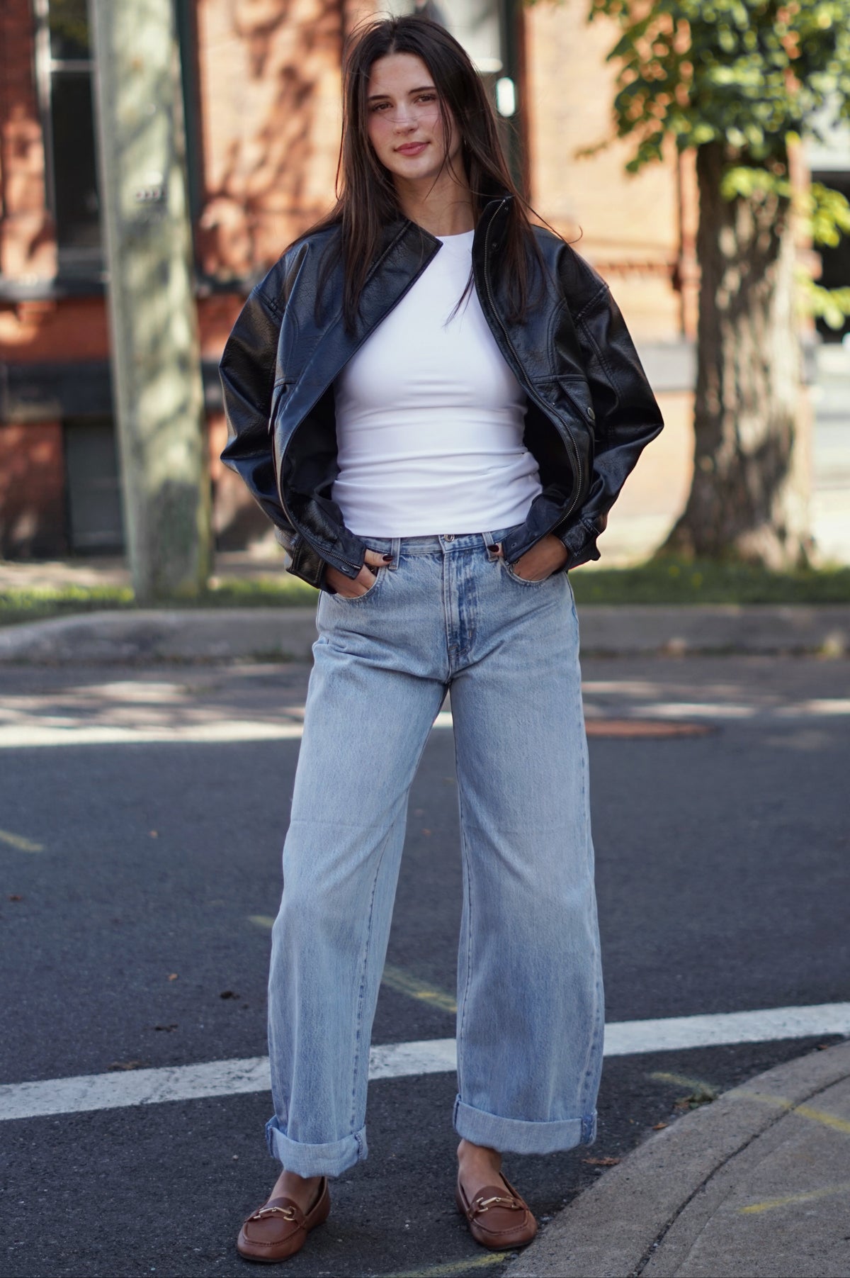 A woman stands on an outdoor street corner wearing a black leather bomber jacket, a white ribbed top, light-wash wide-leg jeans, and brown leather loafers.