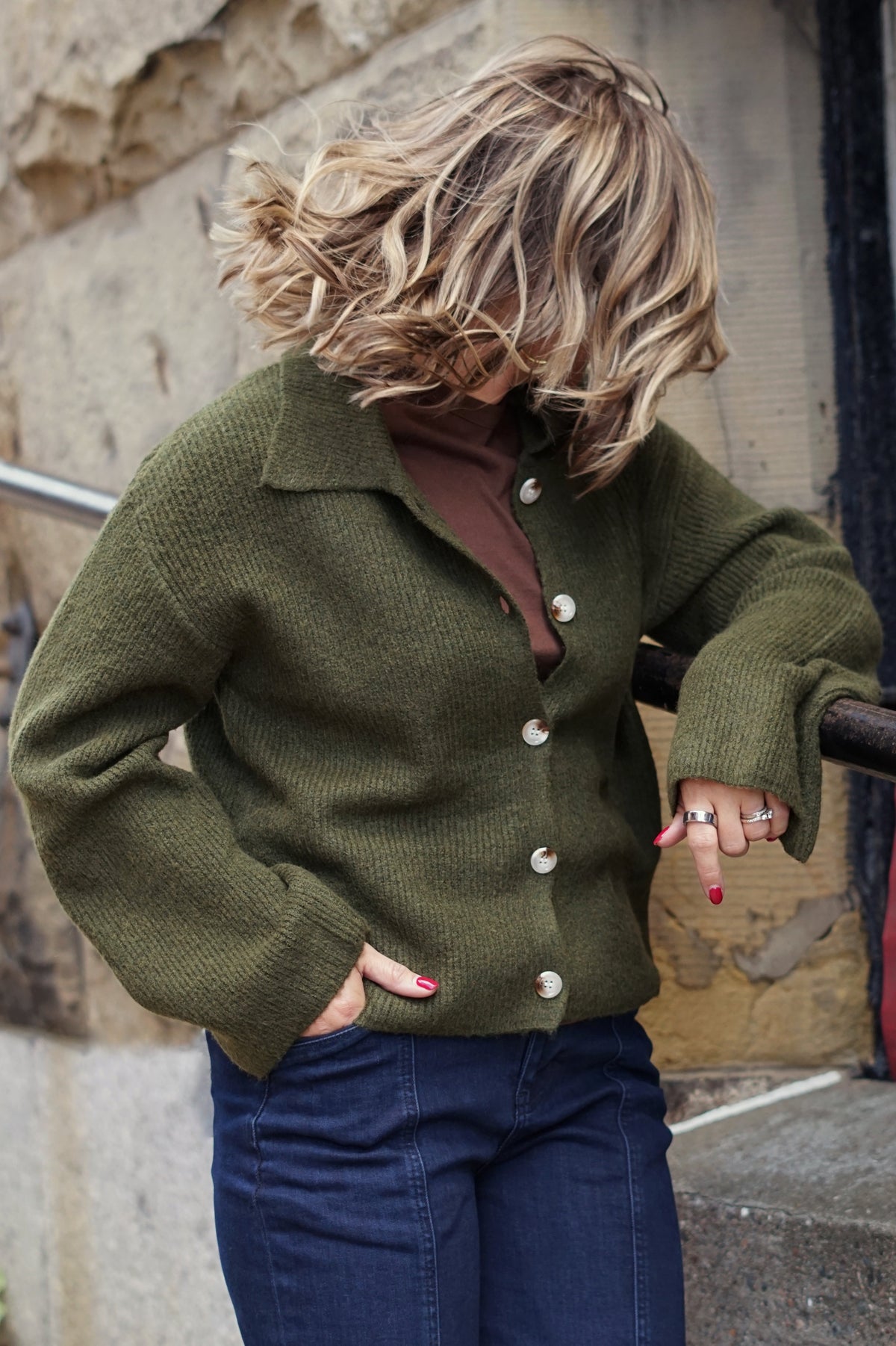 A woman wearing an olive green knit button-up cardigan over a brown top and dark blue jeans, standing outdoors against a stone wall.