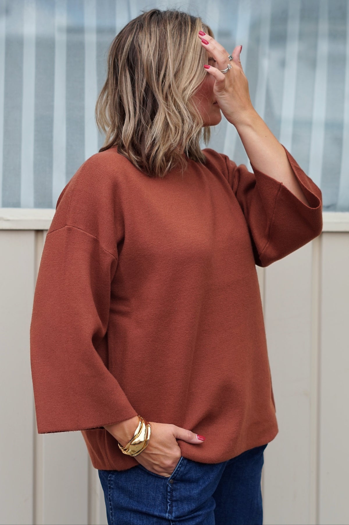 A woman with blonde hair is shown from the back wearing a rust-colored, wide-sleeve knit top and blue jeans