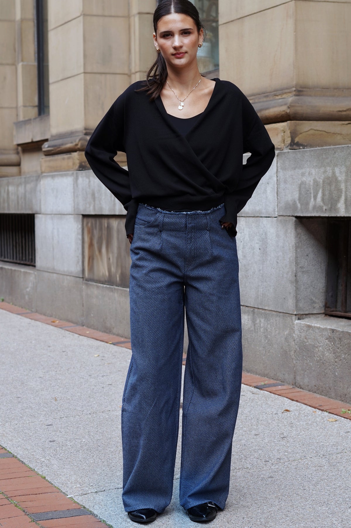 A woman stands on a city sidewalk wearing a black wrap-style long-sleeve top and wide-leg blue jeans, with her hands in her pockets.