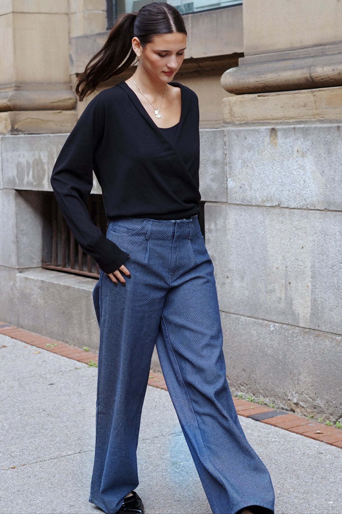 A woman wearing a black long-sleeve top and wide-leg blue jeans stands on a city sidewalk in front of a stone building.