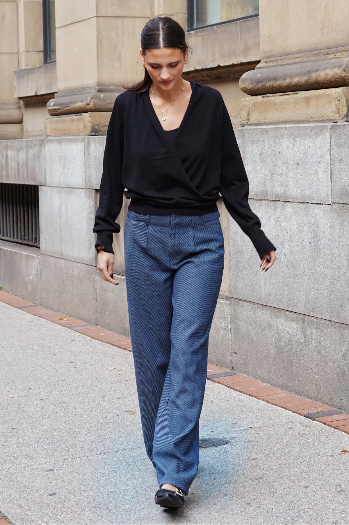 A woman walking outdoors on a city sidewalk next to a stone building, wearing a black long-sleeve wrap-style top and blue wide-leg jeans.