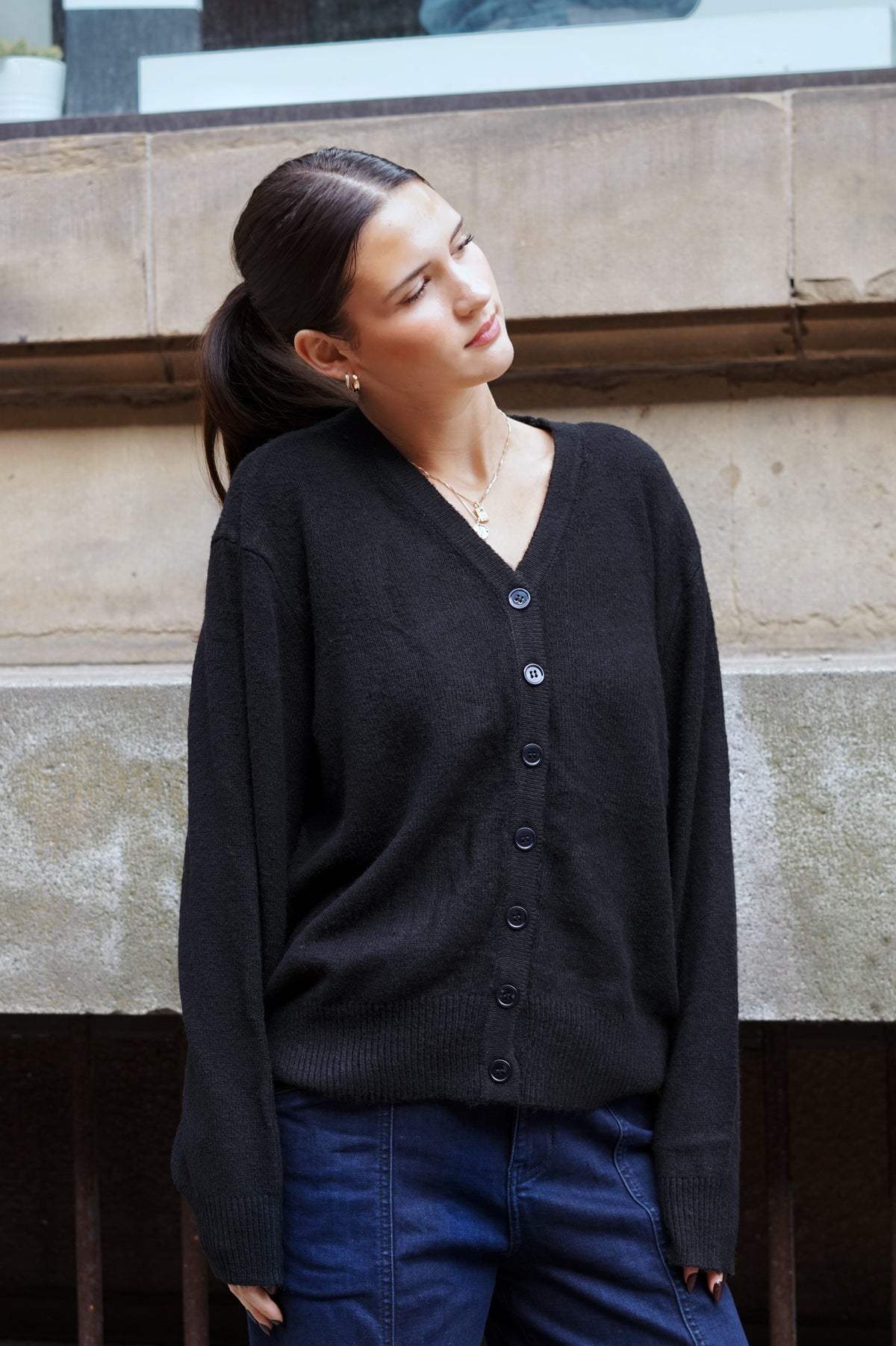 A woman wearing a dark charcoal gray V-neck button-up cardigan sweater and blue jeans, standing against a stone wall.
