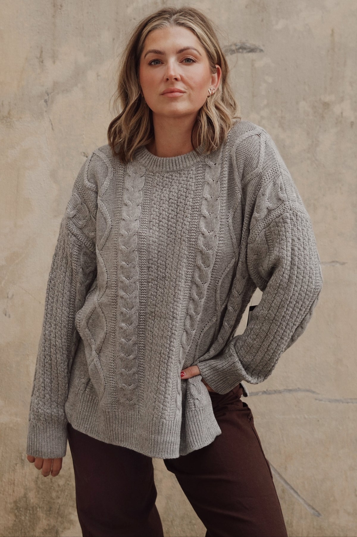 A woman wearing an oversized light gray cable-knit sweater and dark brown pants, standing against a textured beige concrete wall.
