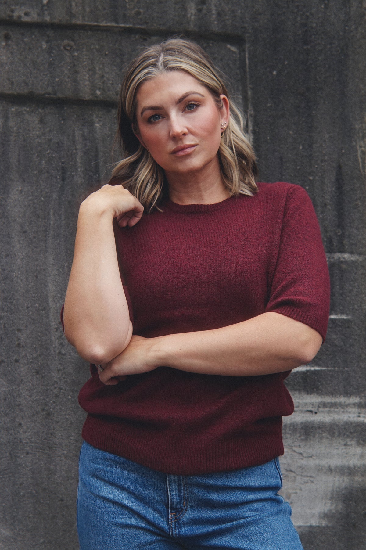 A woman with blonde hair wearing a short-sleeve crewneck knit sweater in burgundy and blue jeans, posing against a dark textured concrete wall.