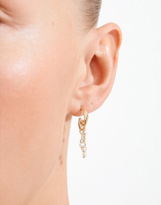 A close-up of a model's ear wearing a delicate gold huggie hoop earring with a dangling charm featuring small clear stones.