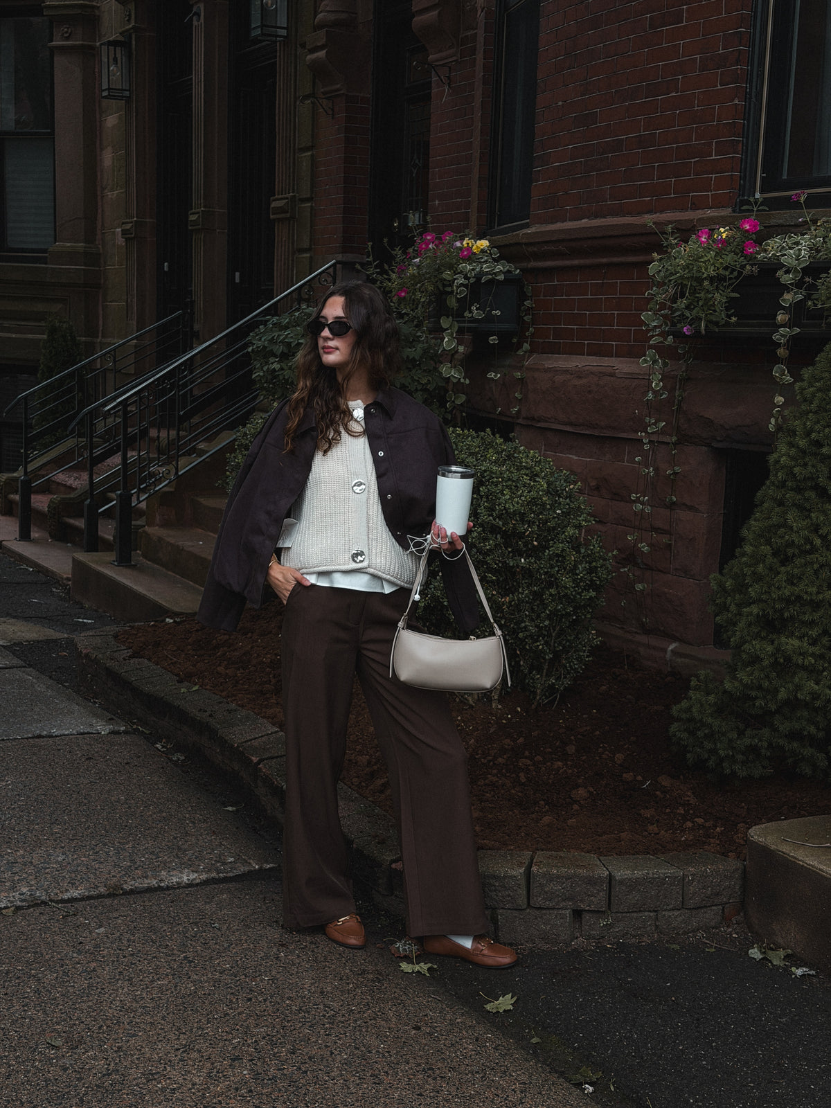 A woman stands on a city sidewalk in front of a brick building, wearing a dark blazer, cream knit top, brown wide-leg pants, and brown loafers. She carries a light beige shoulder bag and holds a takeaway coffee cup.