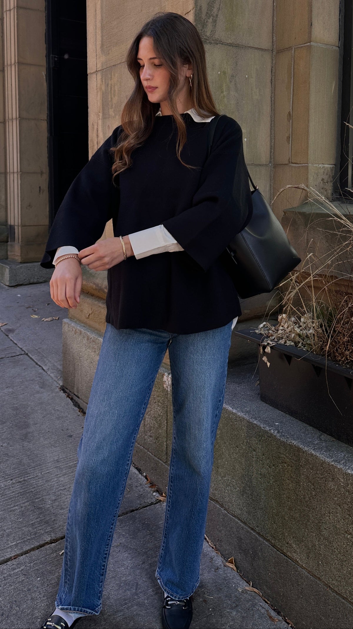 Woman wearing a black wide-sleeve sweater layered over a white collared shirt with blue jeans and a black shoulder bag, standing outside beside a stone building.