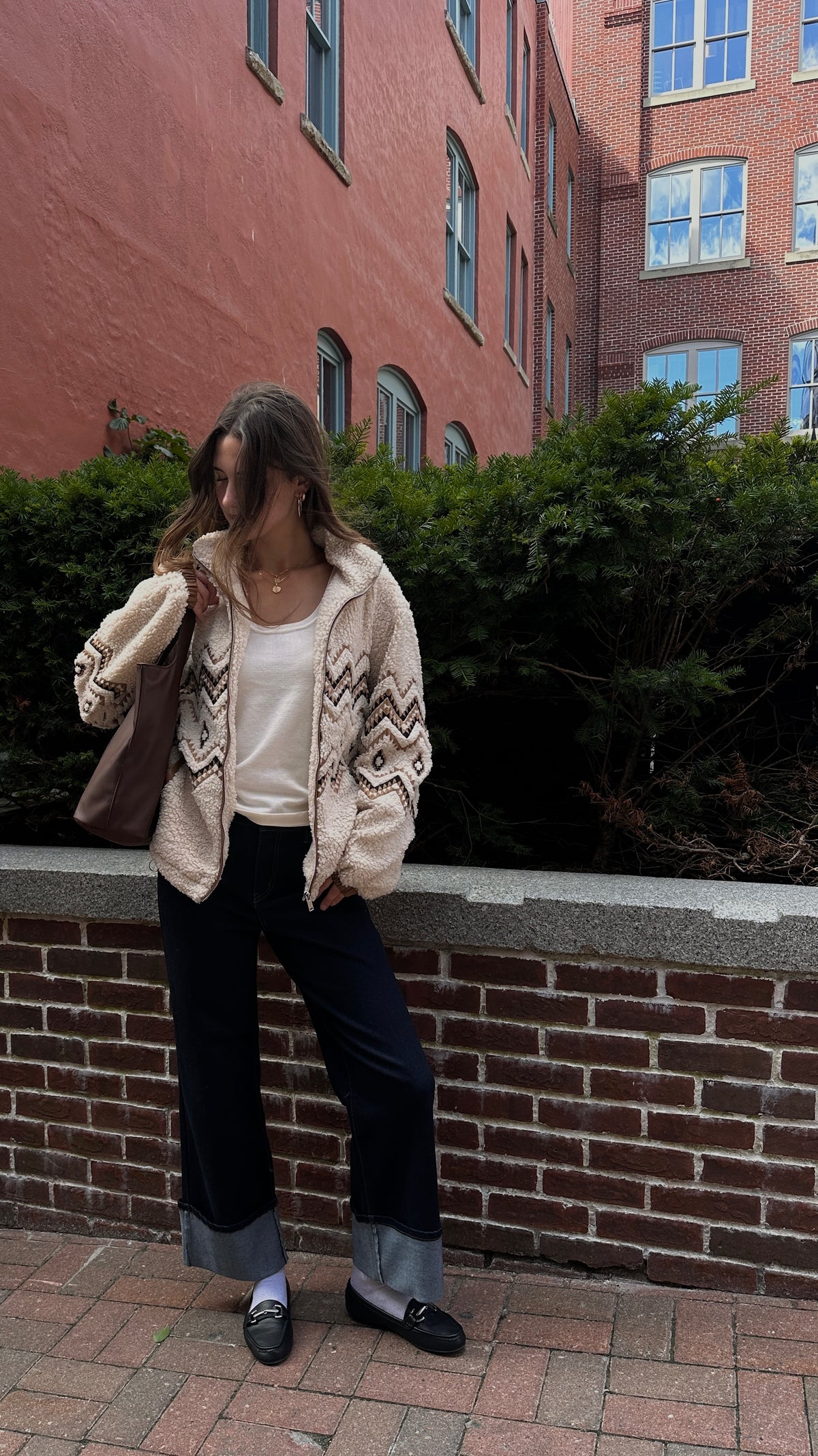 Woman wearing a cream patterned sherpa jacket over a white top with dark cuffed jeans and black loafers, standing outdoors beside a brick wall and greenery.