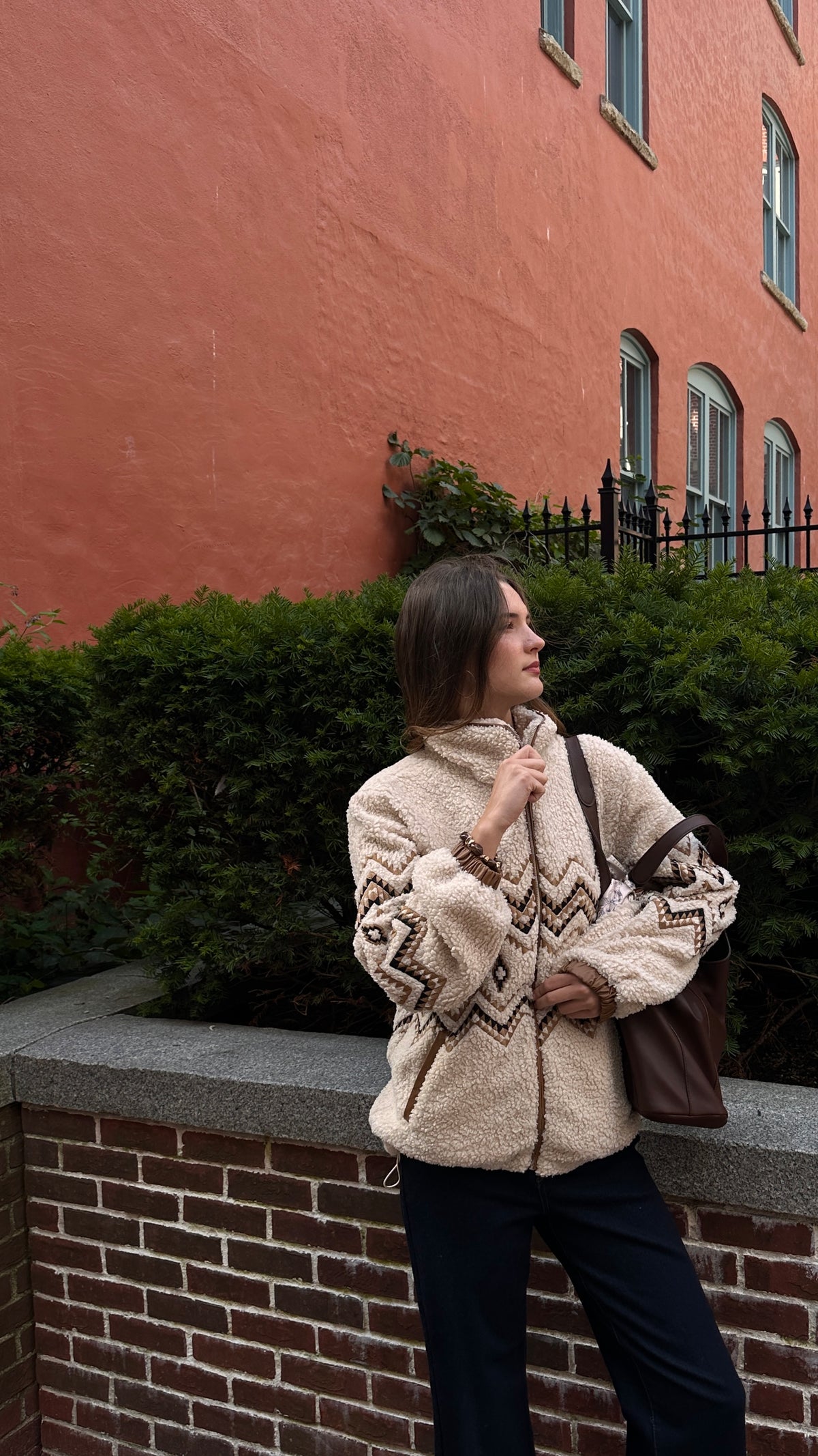 Woman wearing a cream sherpa jacket with brown geometric detailing, dark wide-leg jeans and a brown shoulder bag, standing outdoors beside a brick wall and greenery.
