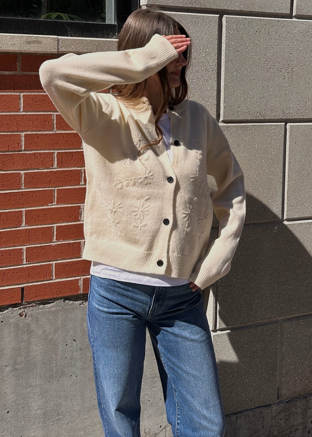 Woman wearing a cream embroidered cardigan layered over a white top with blue jeans, standing outside against a brick and stone building.