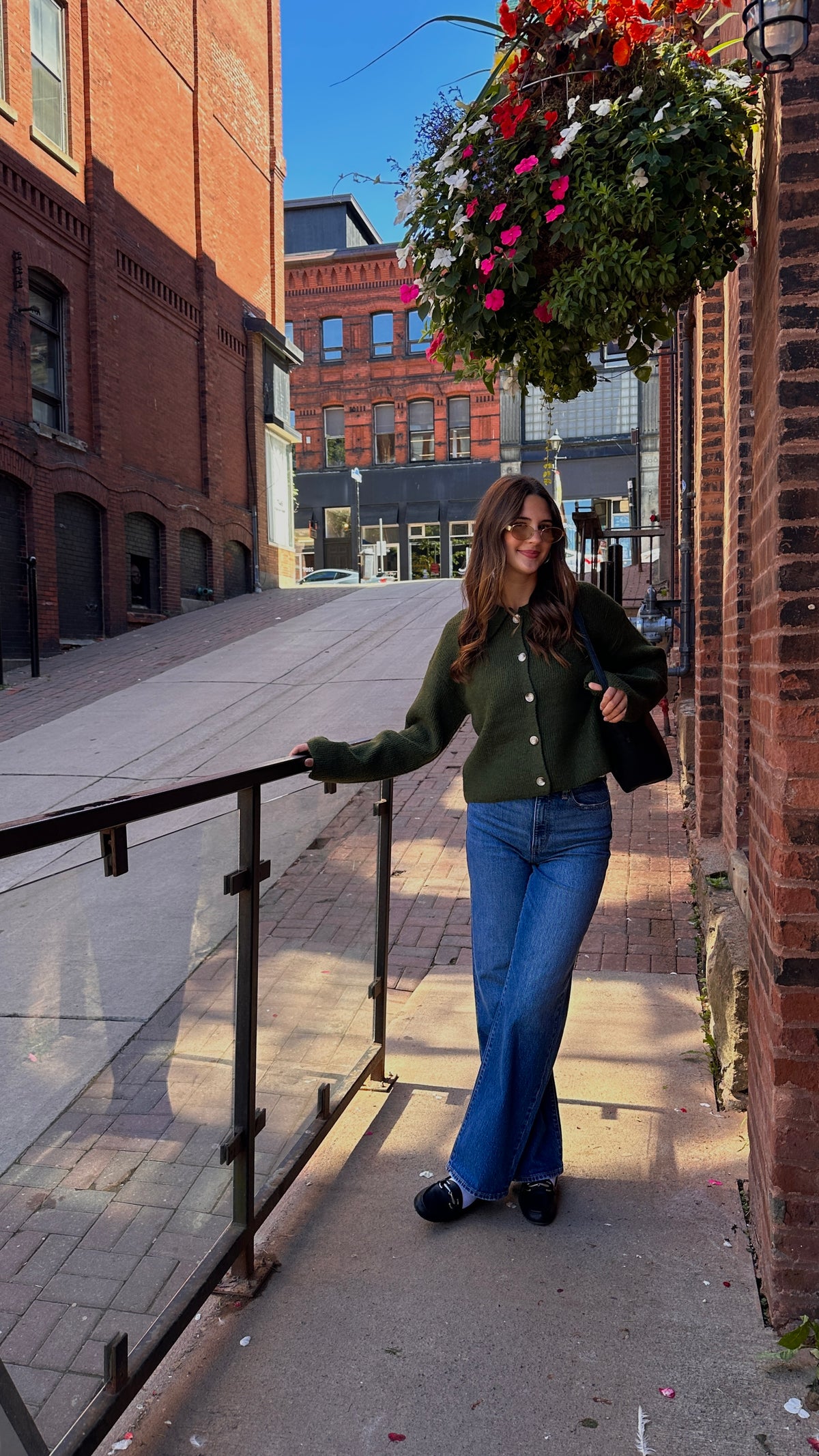 Woman wearing a dark green button-front cardigan with blue jeans, sunglasses and a black shoulder bag on a brick-lined city street.