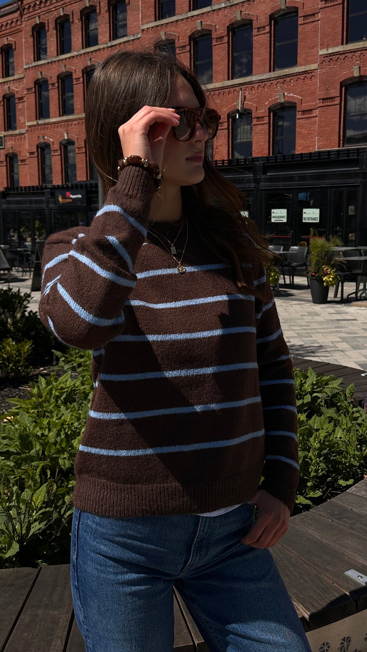 Woman wearing a brown sweater with light blue stripes and blue jeans outdoors in an urban setting.