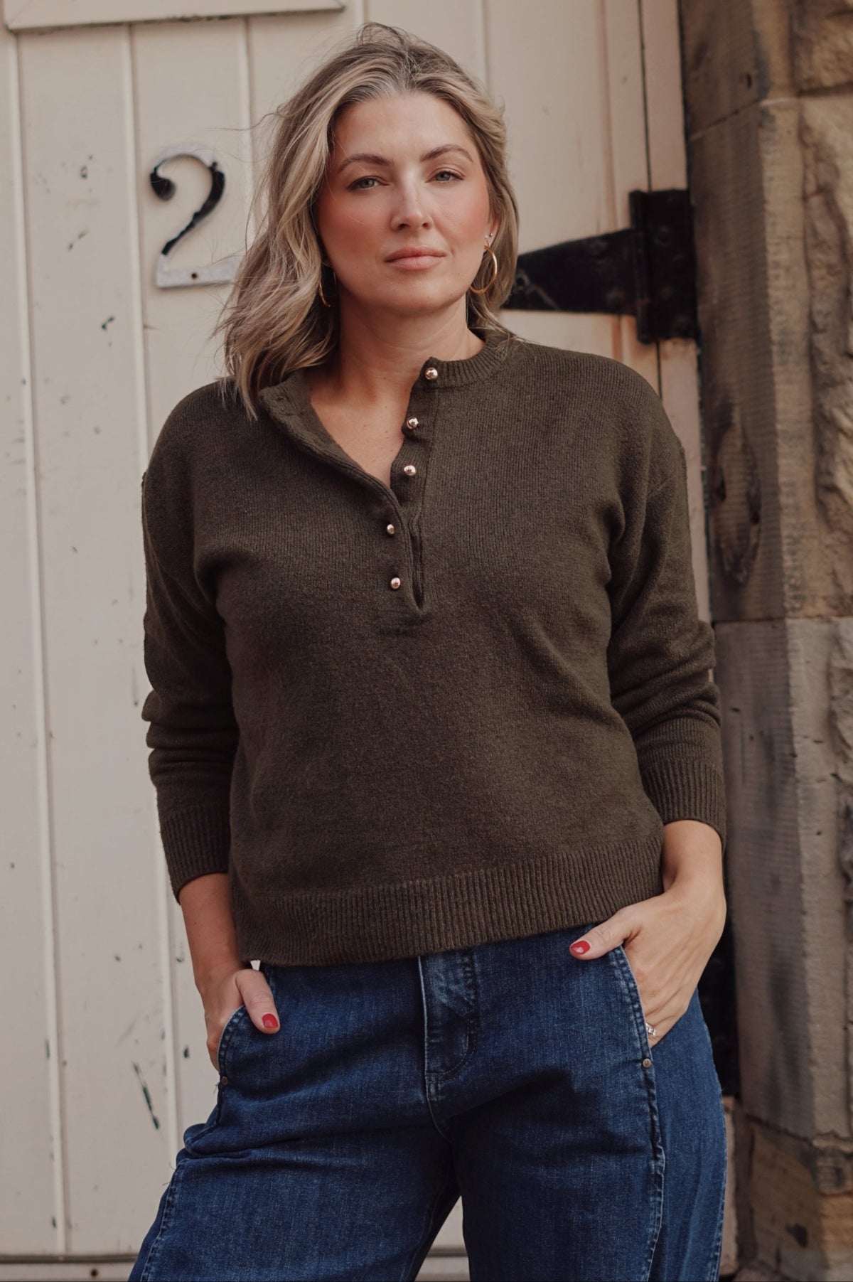 A woman wearing a dark olive brown henley sweater and blue jeans, standing in front of a white wooden door with a stone wall on the right.