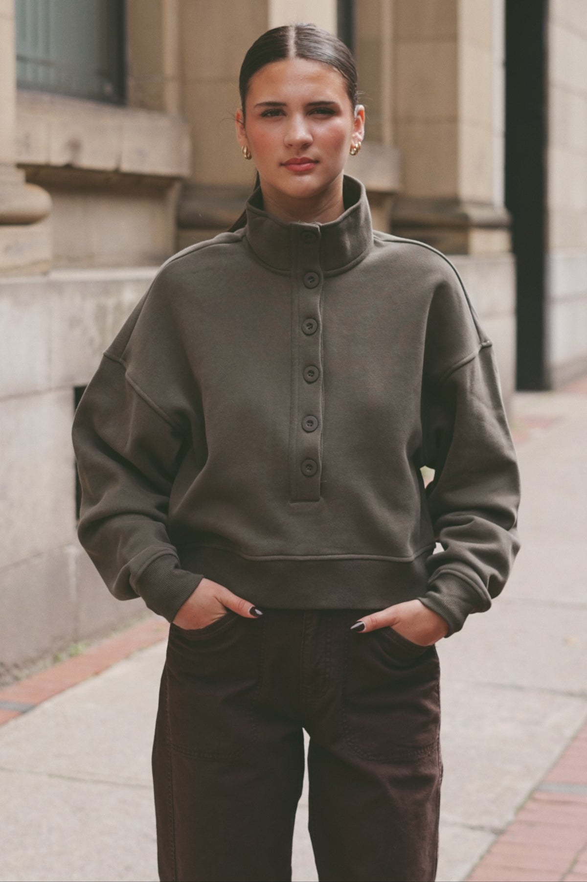 A woman wearing an oversized dark olive green mock-neck button-up sweatshirt and dark brown pants, standing outdoors on a sidewalk in front of a stone building.