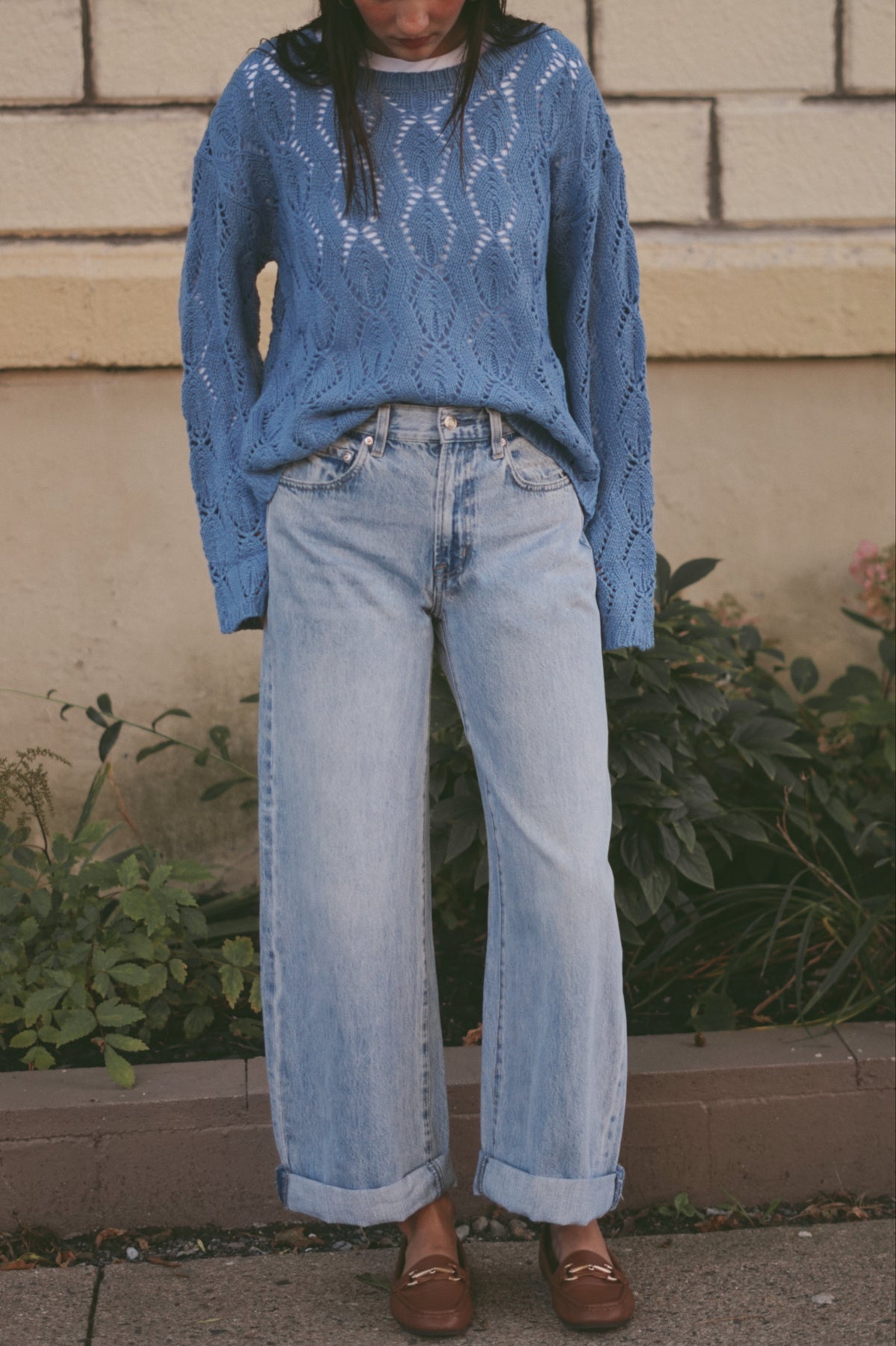 A person wearing a blue open-knit sweater and light-wash wide-leg jeans, standing outdoors in front of a beige brick wall and greenery.