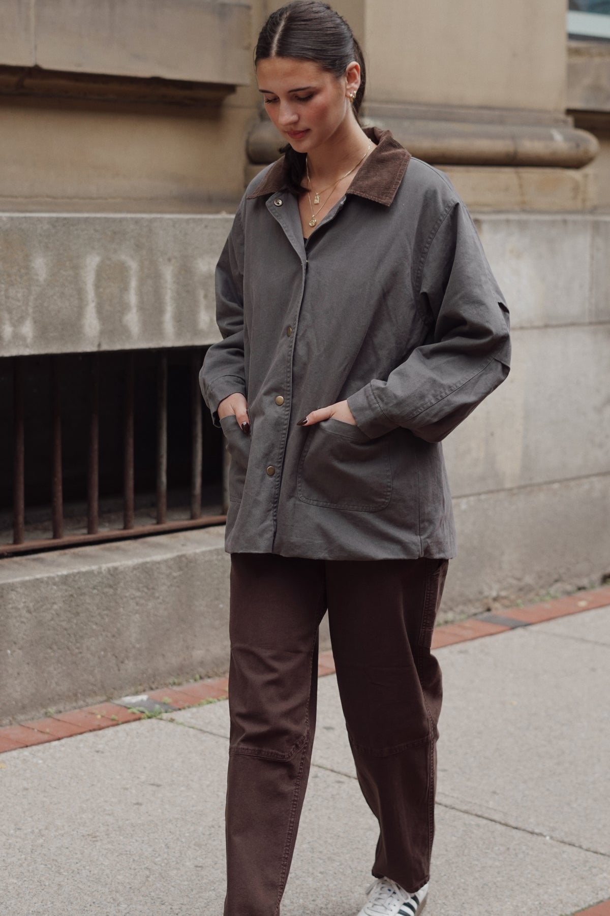 A woman wearing a dark gray waxed cotton utility jacket with a brown corduroy collar, paired with brown pants, standing on a city sidewalk.