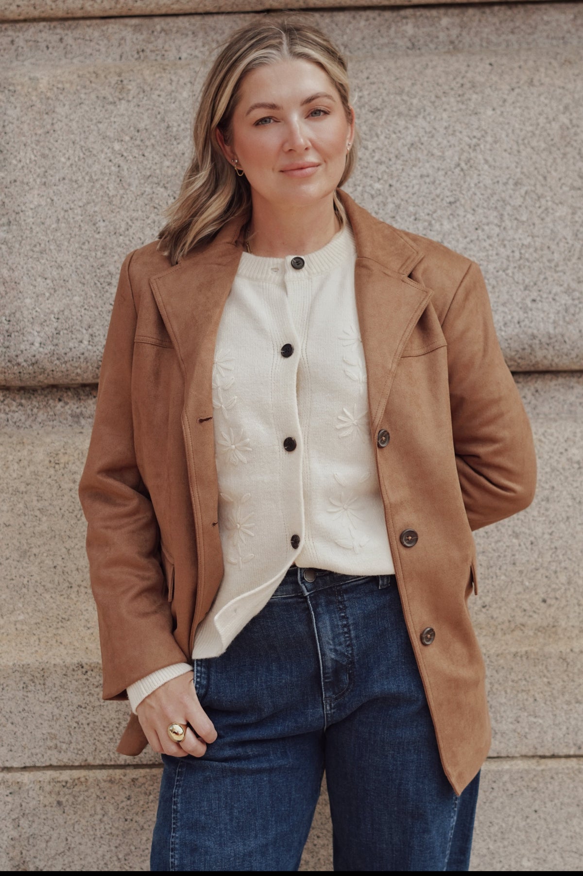 A woman wearing a camel brown suede blazer over a cream button-up shirt and blue jeans