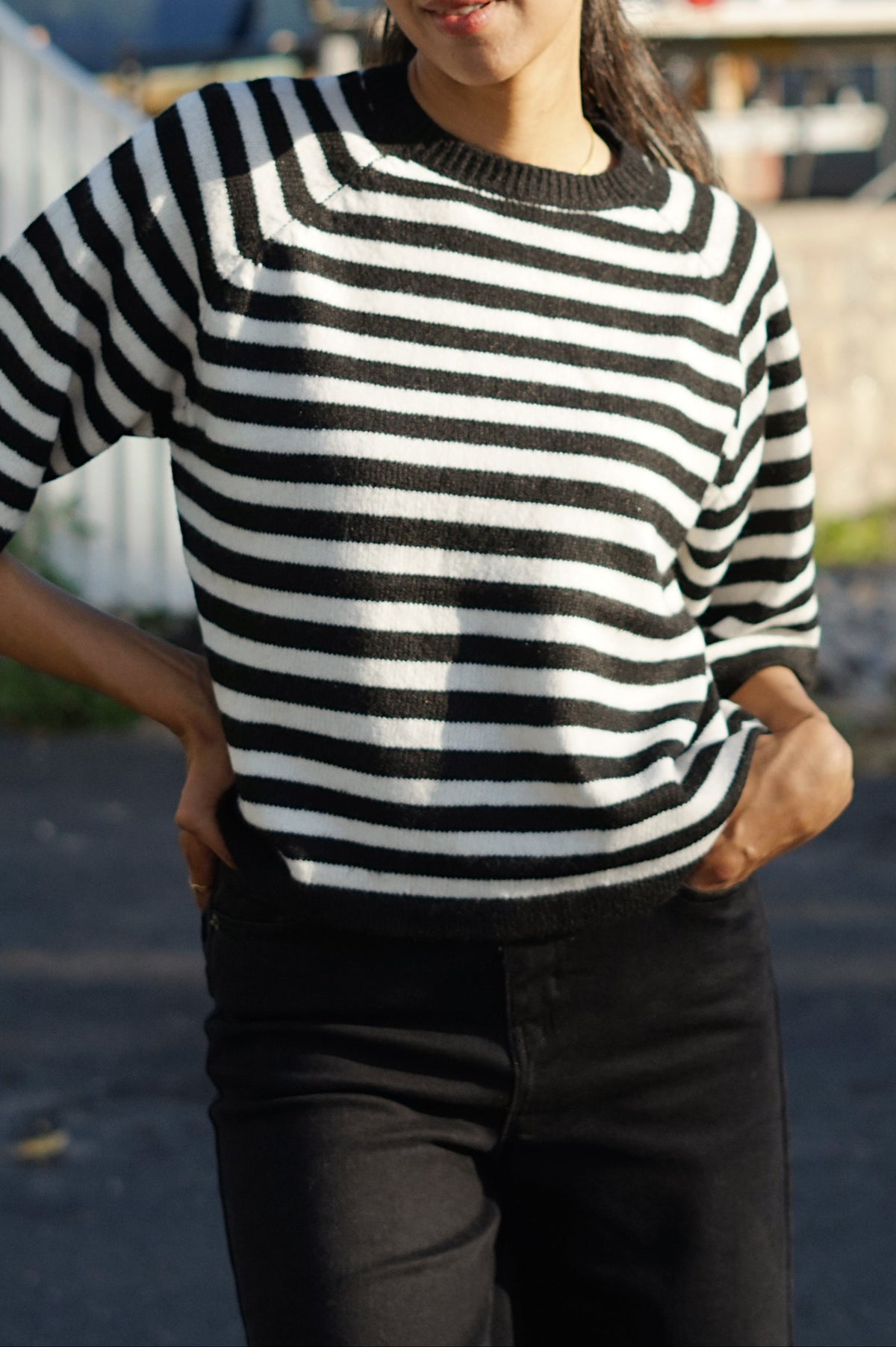 A woman wearing a black and white horizontally striped short-sleeve knit top and black pants, standing outdoors with her hands on her hips in front of a boat and trees.