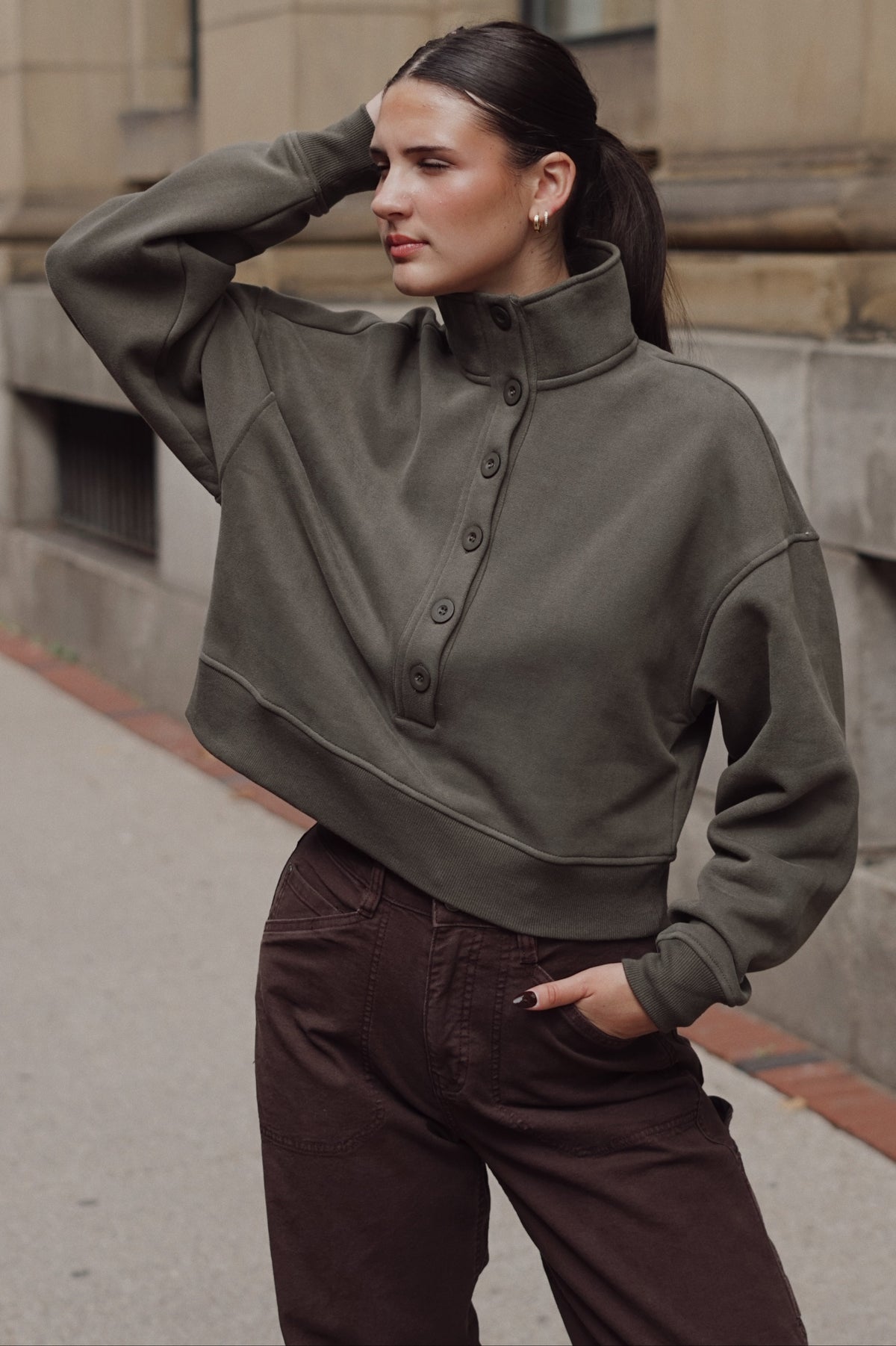 A woman wearing an olive green cropped button-front sweatshirt and dark brown pants, posing outdoors against a stone building background.