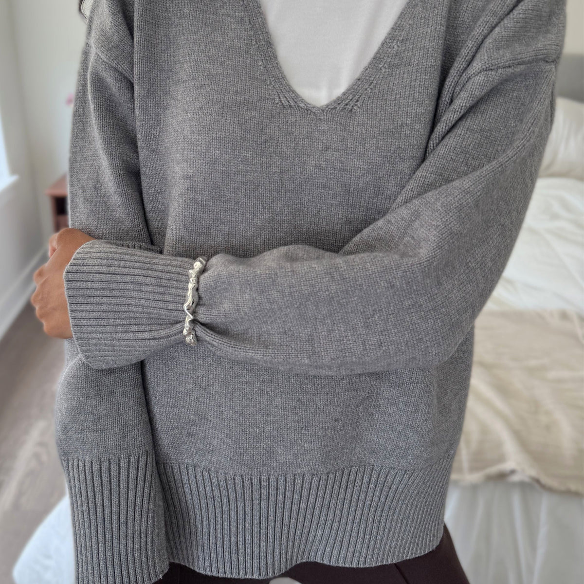 A person wearing an oversized heather gray knit V-neck sweater over a white crewneck shirt, paired with dark leggings, standing in a bedroom setting.