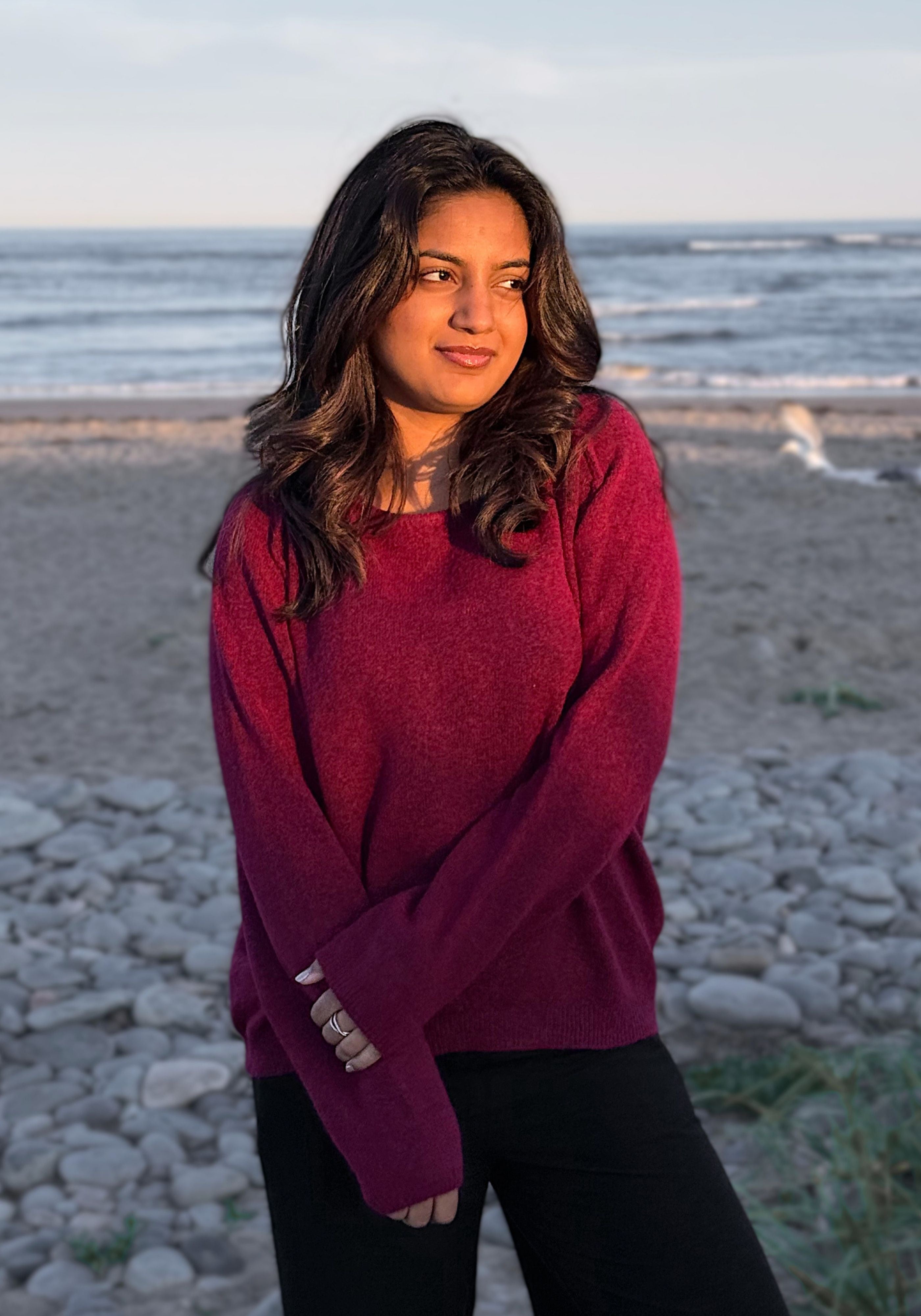A woman wearing a heathered magenta knit sweater and black pants standing on a rocky beach with the ocean in the background.