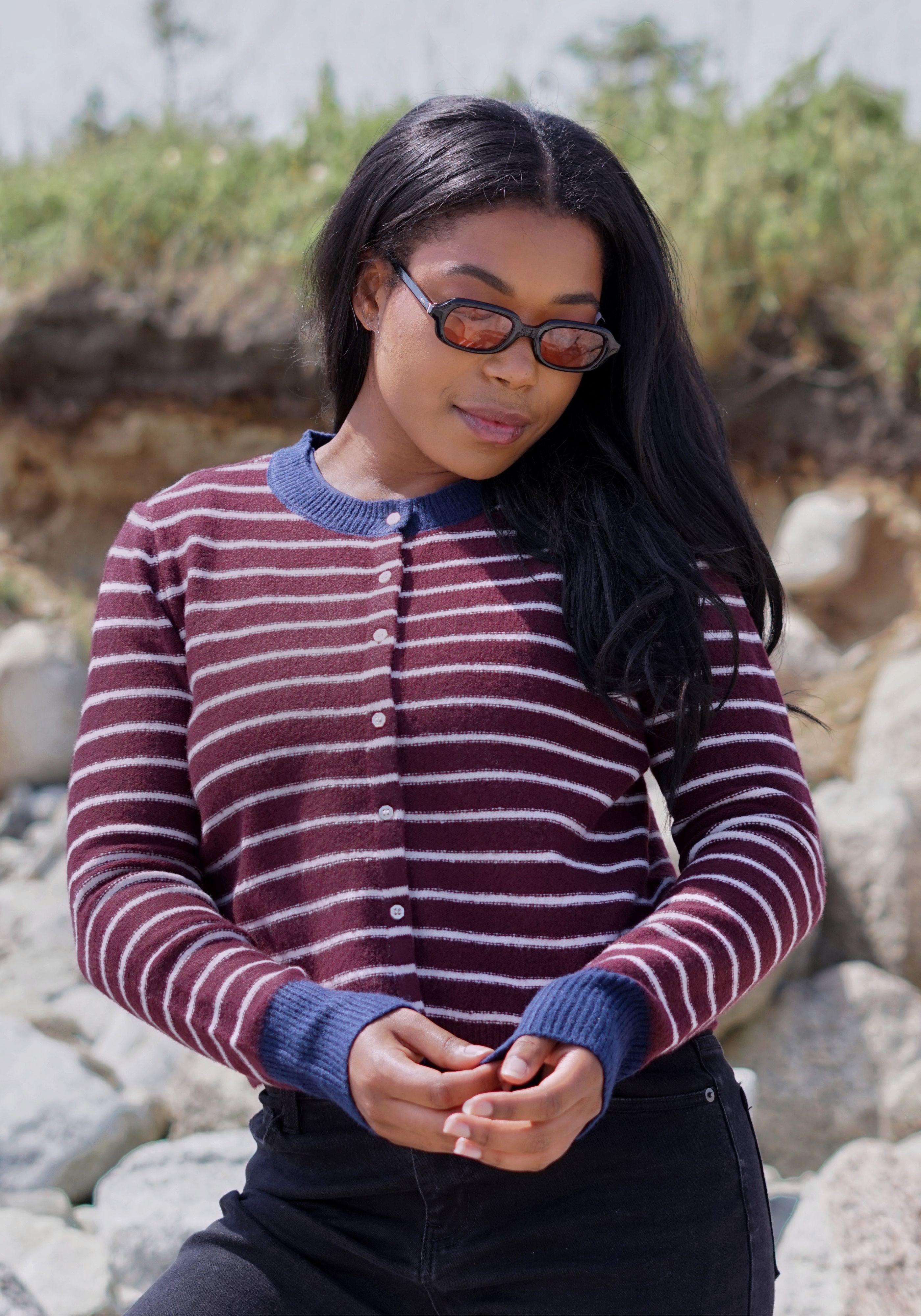 A woman wearing a burgundy and white striped knit cardigan with a navy blue collar, cuffs, and button placket, styled with dark sunglasses and black pants.