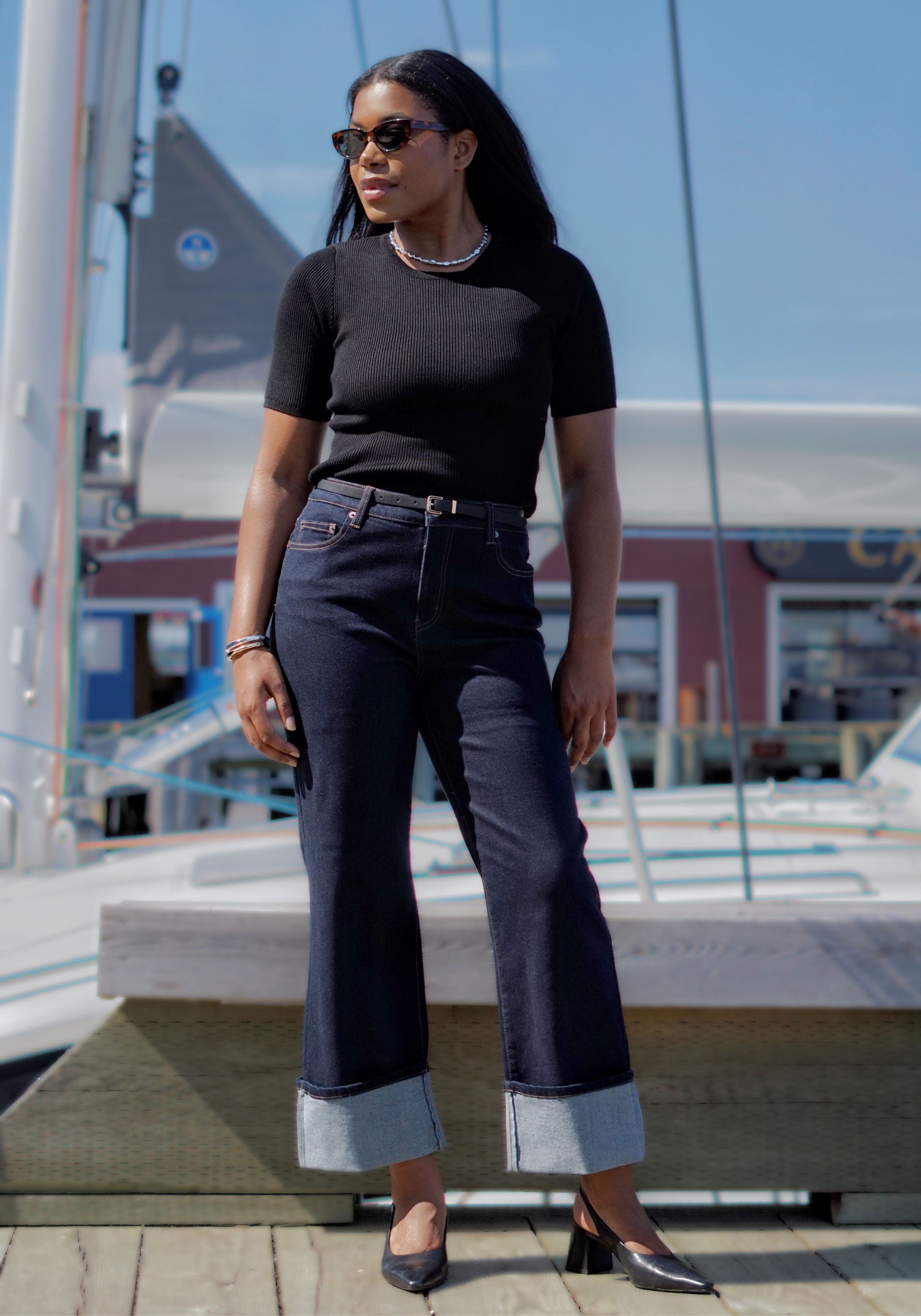 A woman stands outdoors on a dock wearing a fitted black short-sleeve top, dark blue wide-leg jeans with rolled cuffs, black heels, and sunglasses.